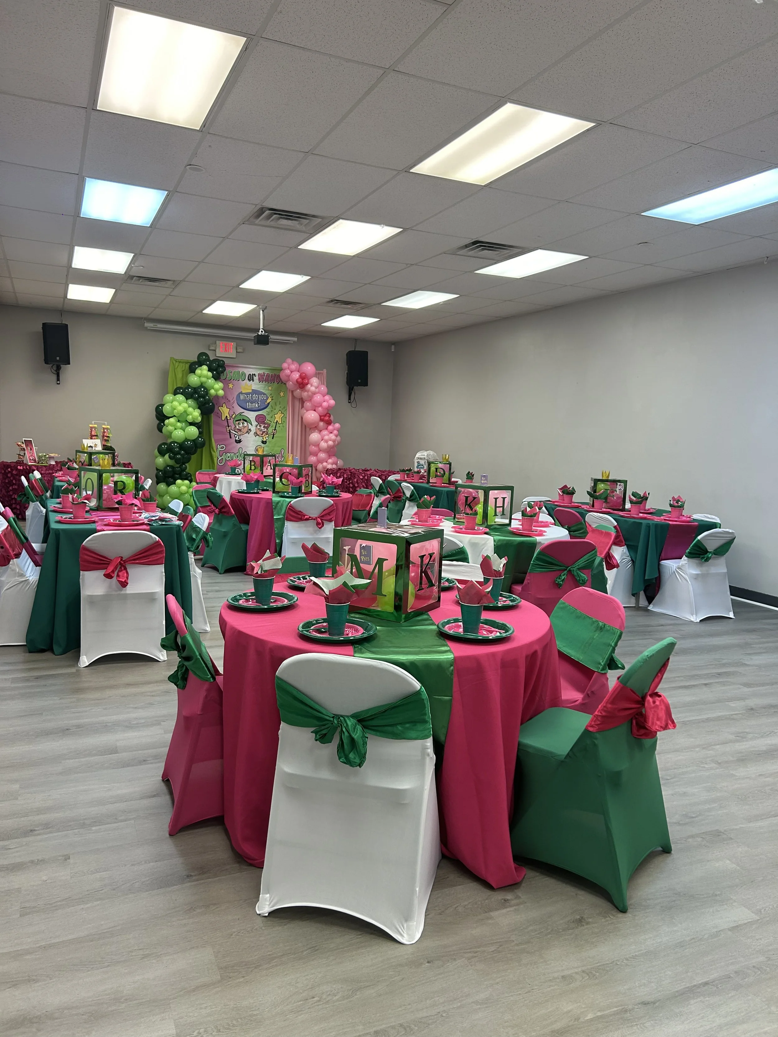 Party room decorated with pink and green themed tablecloths, chairs with pink and green ribbons, and centerpieces. Pink, green, and black balloons form an arch at the back. A colorful backdrop with cartoon characters and the words 'Sao or Whate' is v
