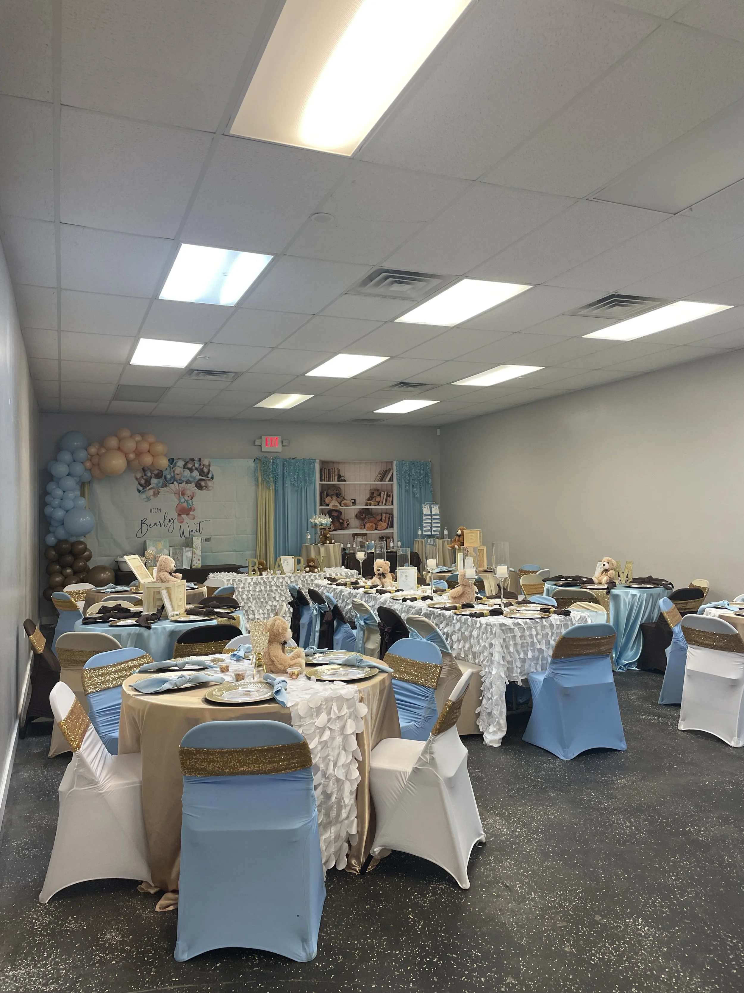 Decorated celebration party room with tables set for guests, teddy bears, balloons, and a backdrop with a book theme for a baby shower.