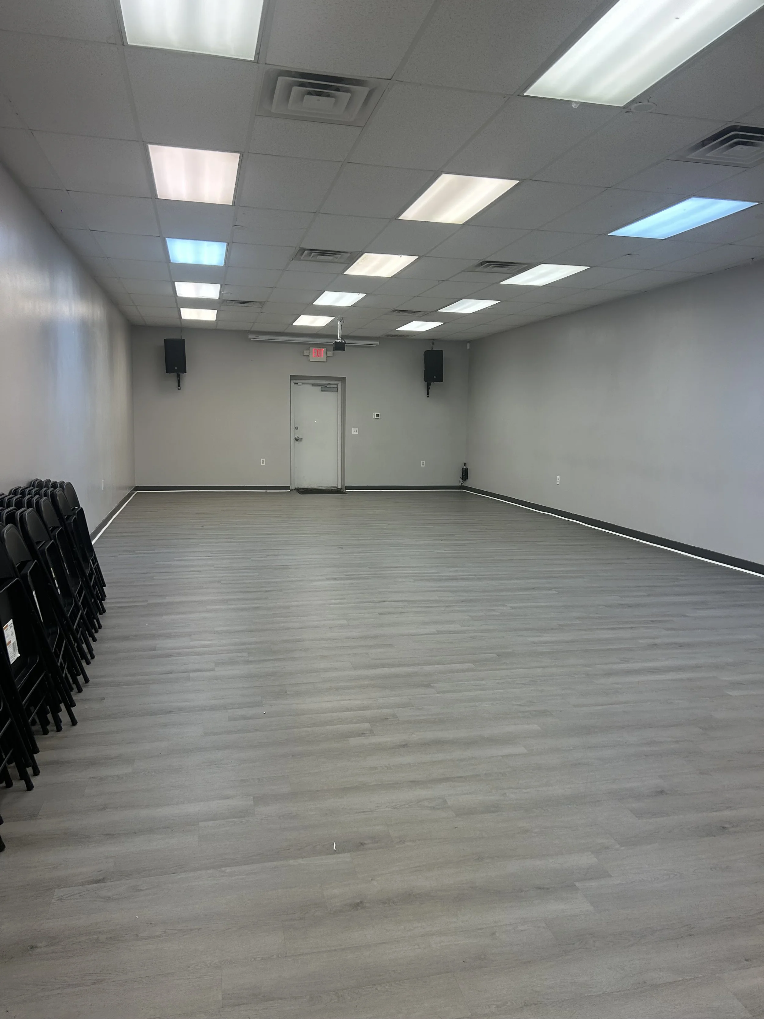 Empty room with wooden floor, white walls, ceiling with fluorescent lights, black stacked chairs on the left, and a door at the back with two speakers on the walls.