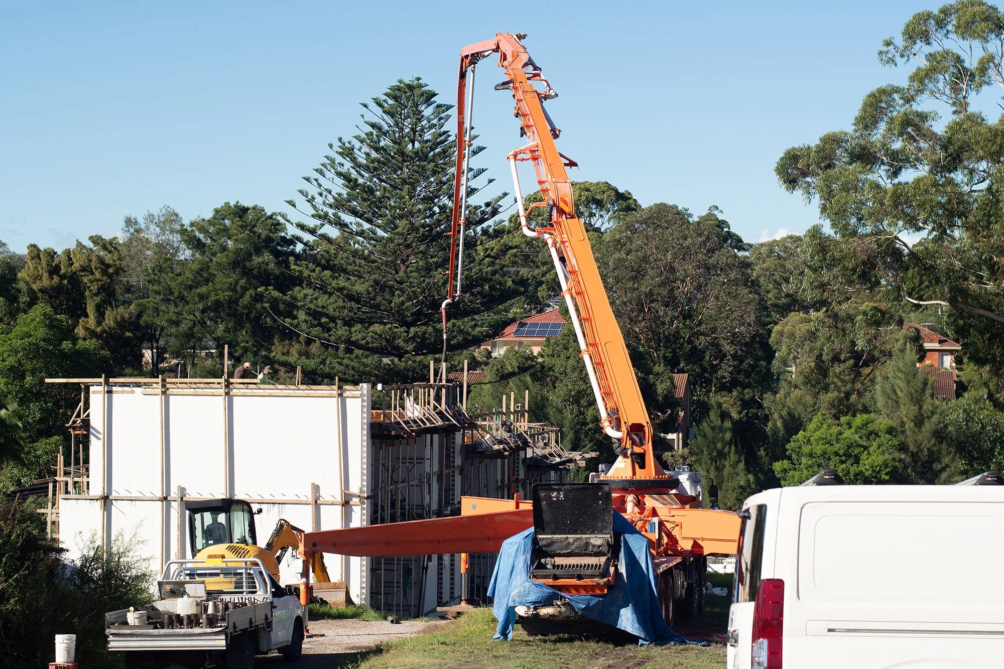 Construction site with a large orange crane lifting materials, a small excavator, and a partially built structure surrounded by trees.