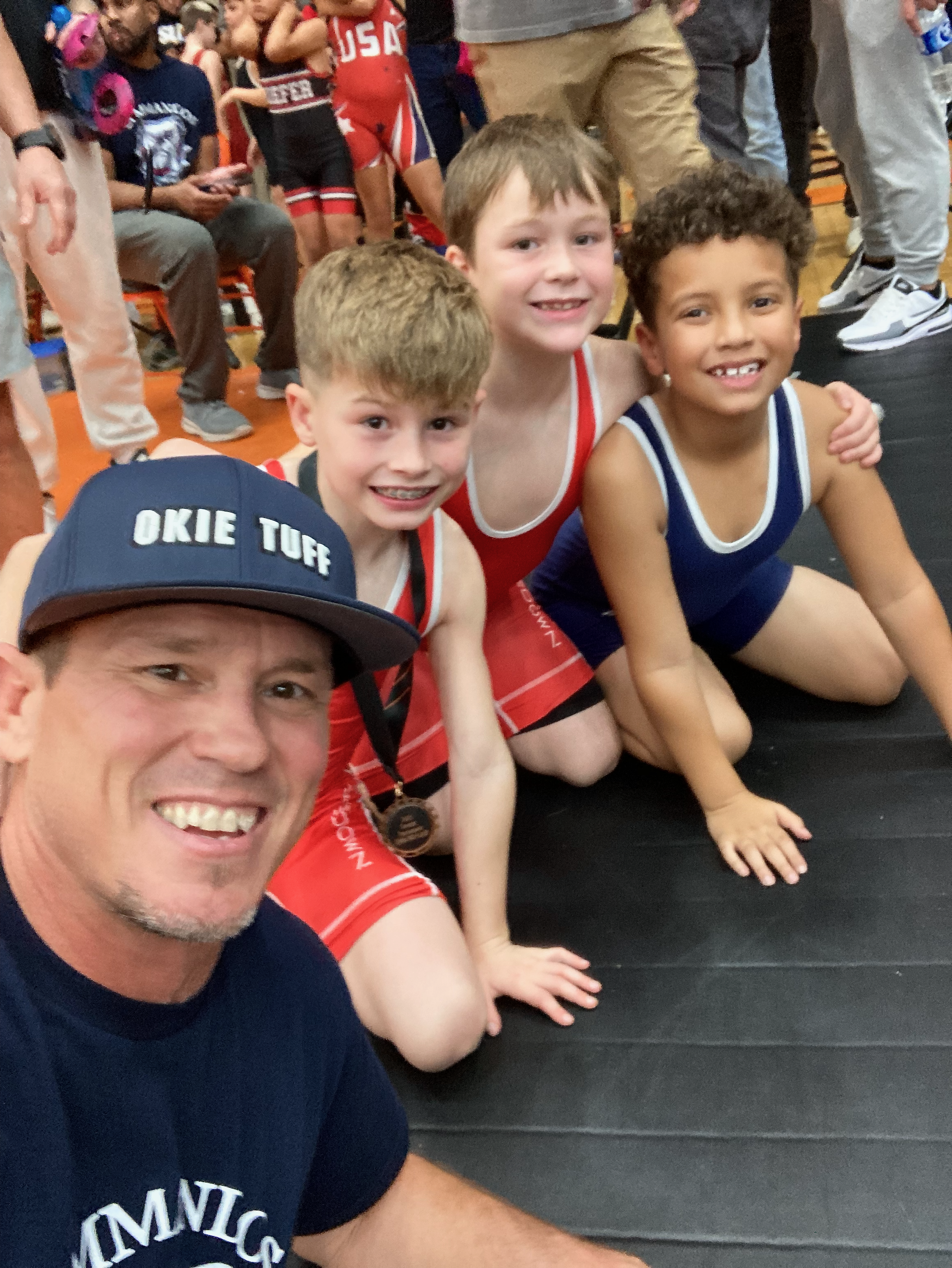 Three young boys, two with curly brown hair and one with straight blond hair, sitting on the floor and smiling at the camera, surrounded by people at a sports event or gathering. One boy is wearing a red and white sports uniform, and another a blue and white sports uniform. The background shows other children and adults, some in athletic attire.