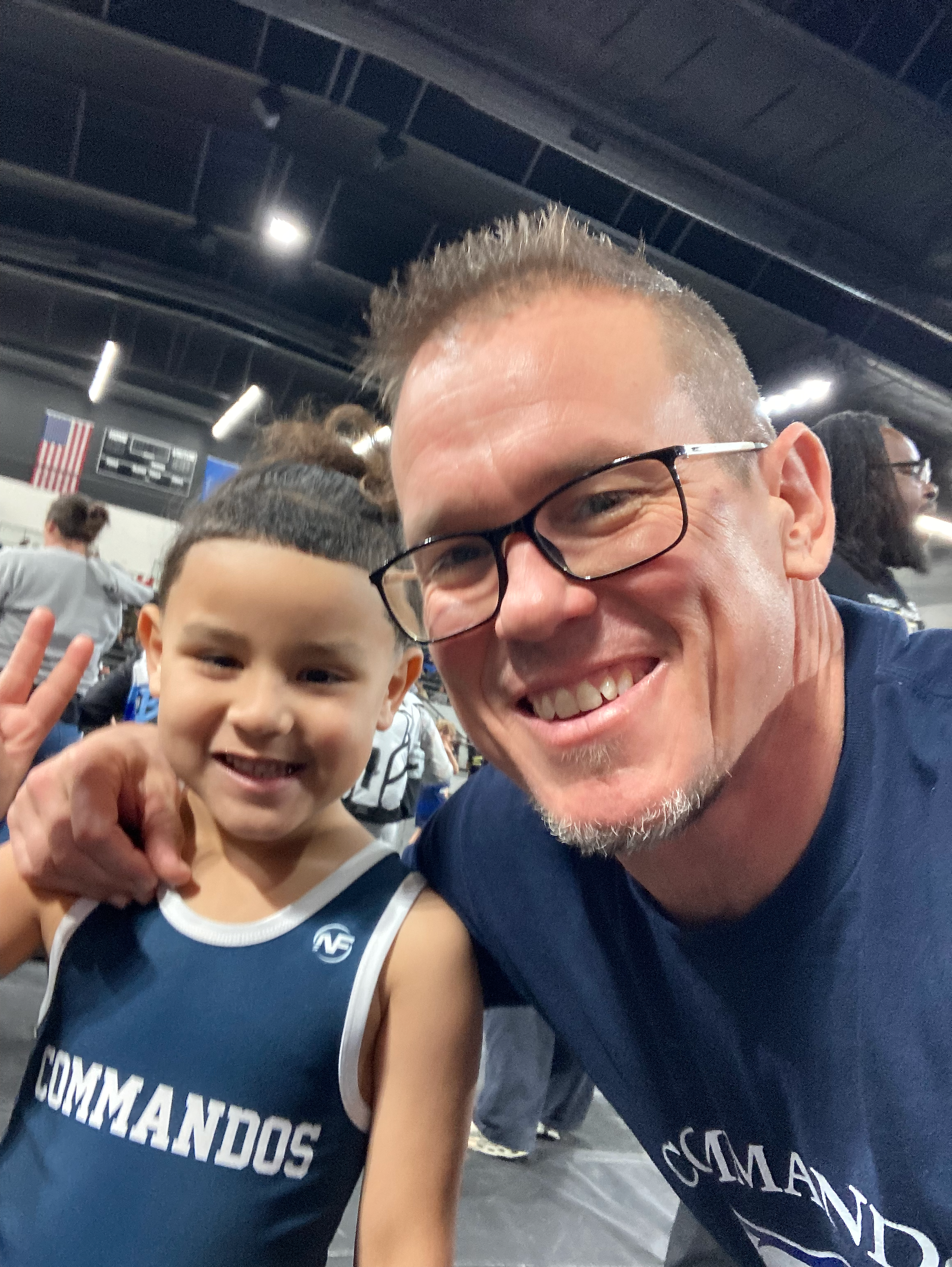 A man with glasses smiling and a young boy wearing a sports jersey, both taking a selfie in what appears to be a gym or sports arena with people in the background.