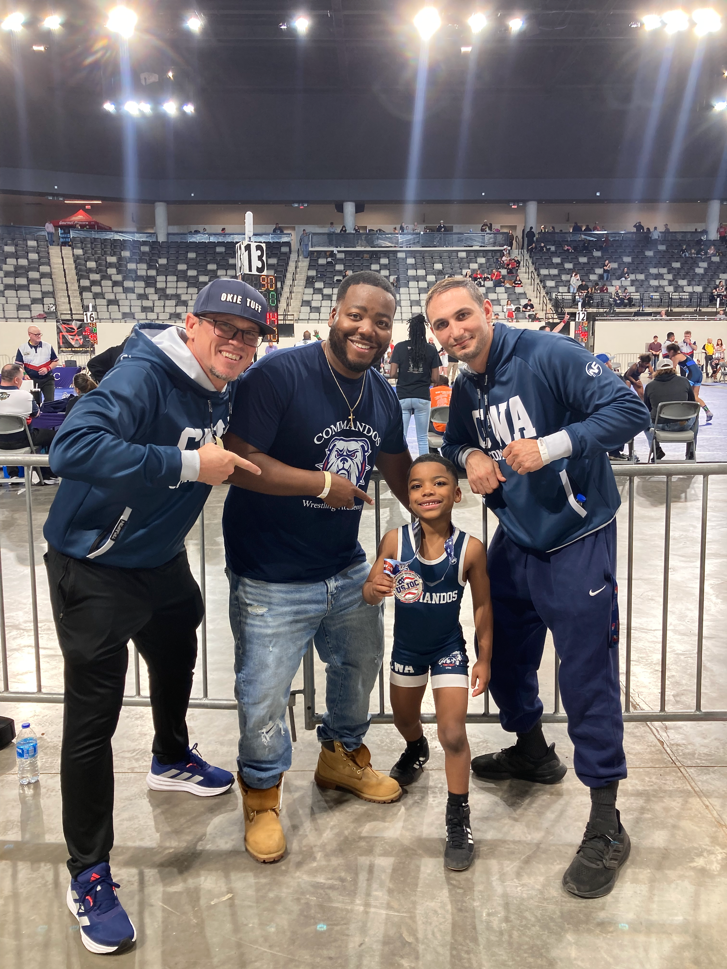 Three men and a young girl are posing together inside a large sports venue with empty seats in the background. The men are dressed in sportswear, and the girl is wearing a sports uniform and holding a medal.