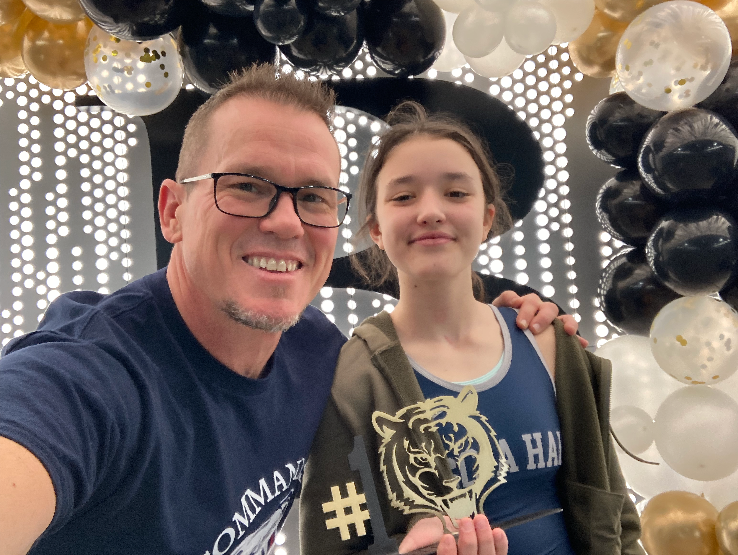 A man with glasses and a young girl are taking a selfie at an event decorated with balloons and lights. Both are smiling and wearing matching dark blue shirts with a lion logo.
