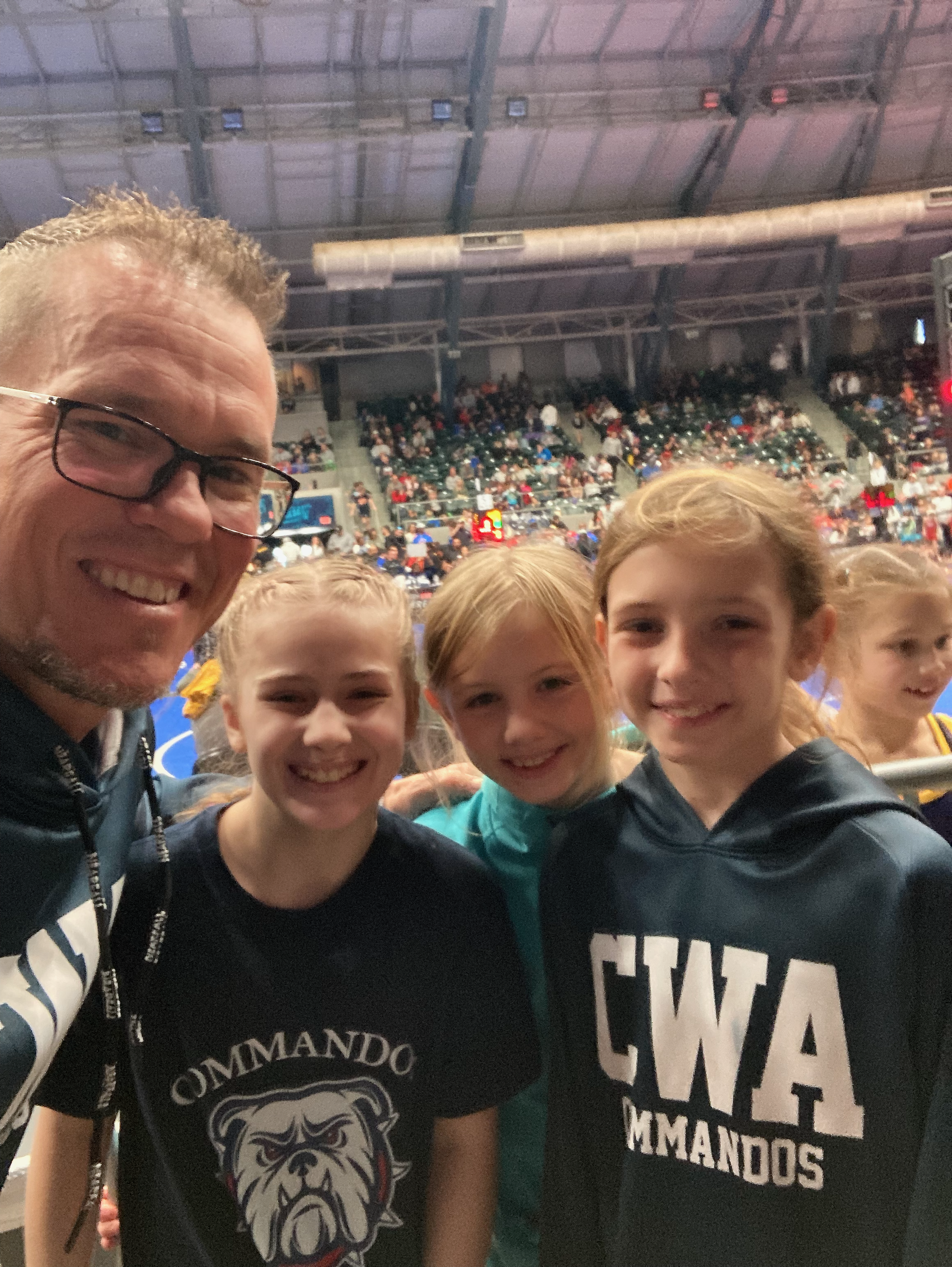 A group of people at an indoor sports event, including a man with glasses smiling and children in front of him, with a crowded stadium seating in the background.