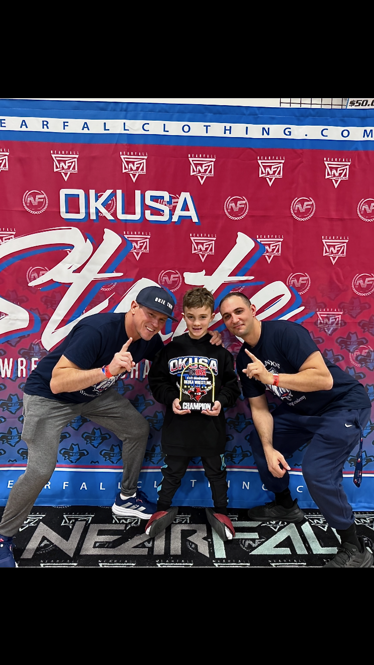 A young boy holding a championship plaque with two men kneeling on either side, all smiling, in front of a red and blue backdrop with the logos 'OKUSA' and 'NEARFALL CLOTHING.'