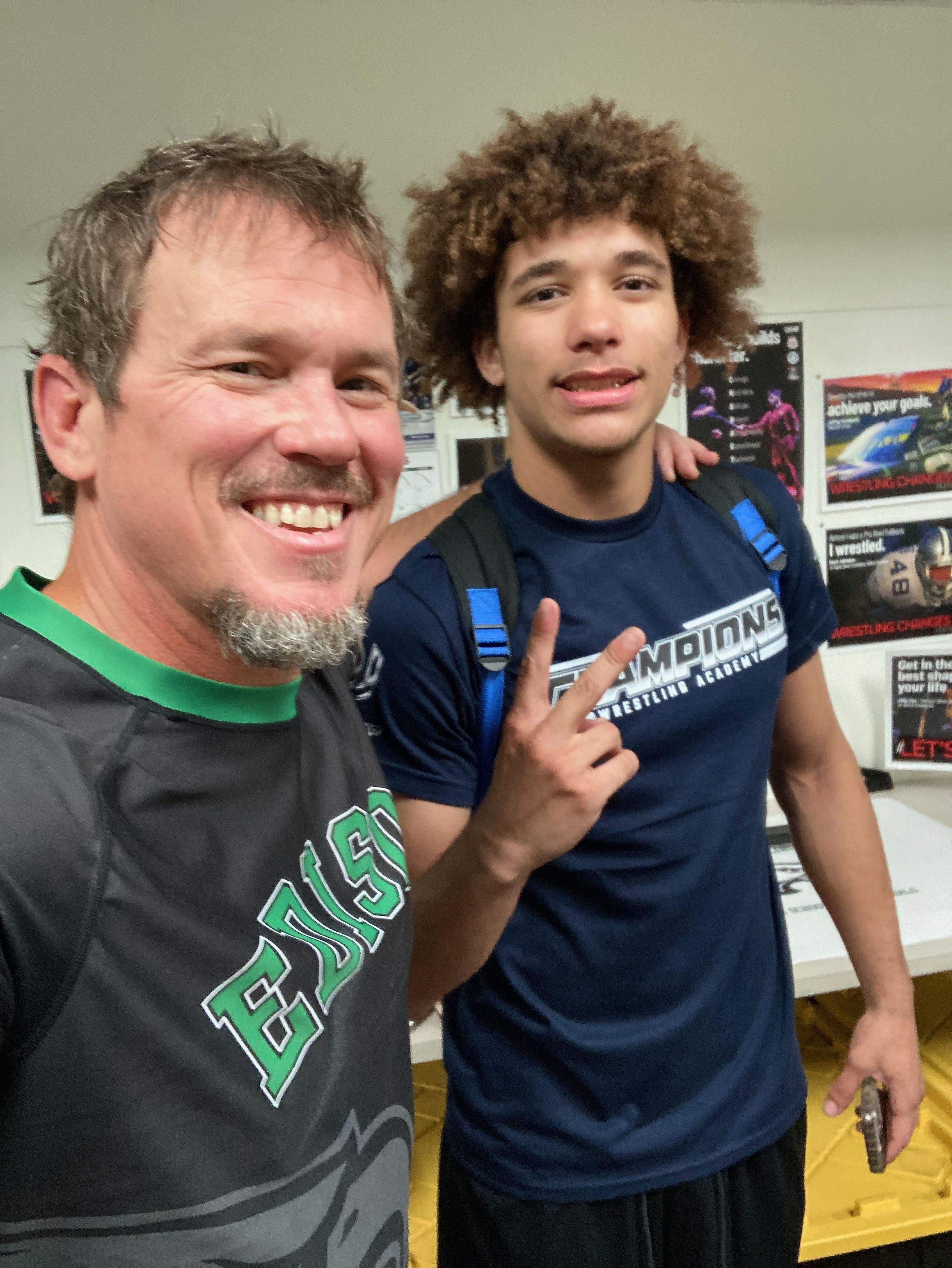 A smiling middle-aged man with a beard wearing a black sports shirt with green accents and a young man with curly brown hair wearing a blue t-shirt with text, standing close together indoors. The background shows posters on the wall and a classroom setting.