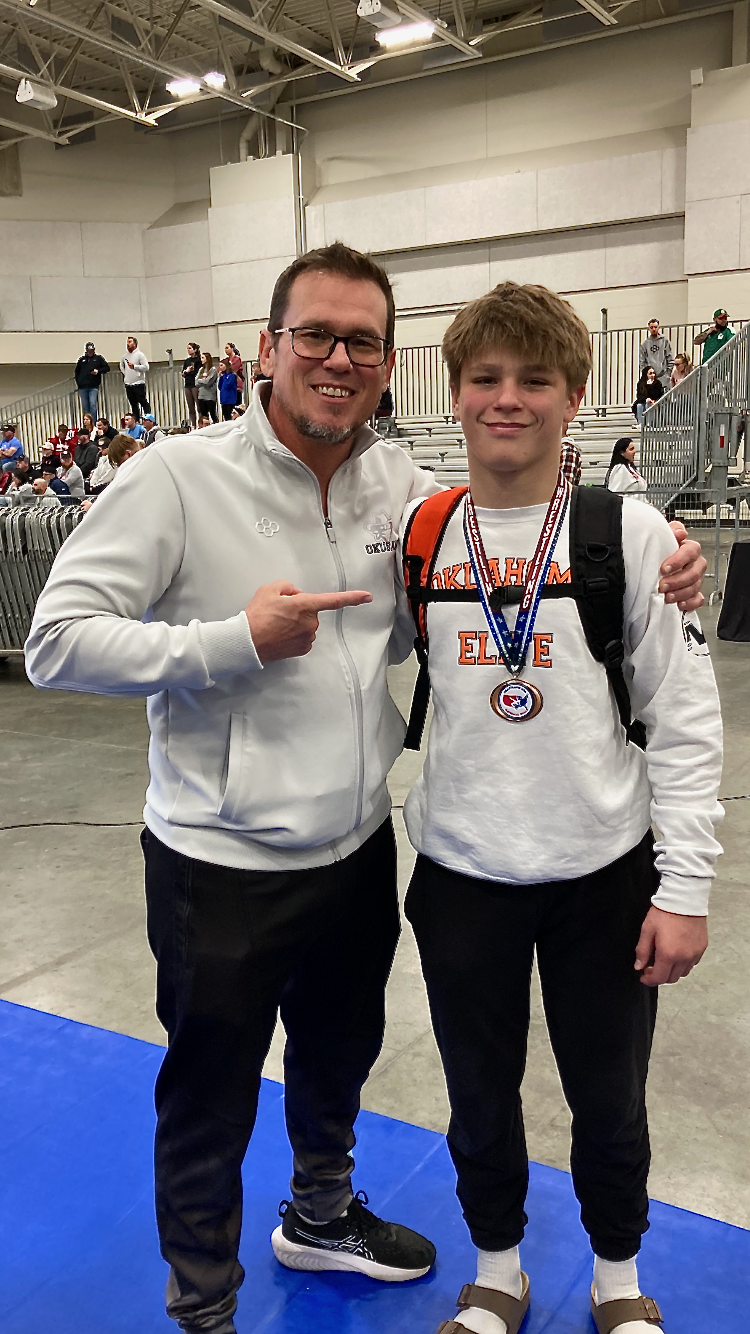 An adult and a teenage boy standing together at an indoor sports event, with the boy wearing a medal around his neck and a backpack. The adult is pointing at the boy, both smiling.