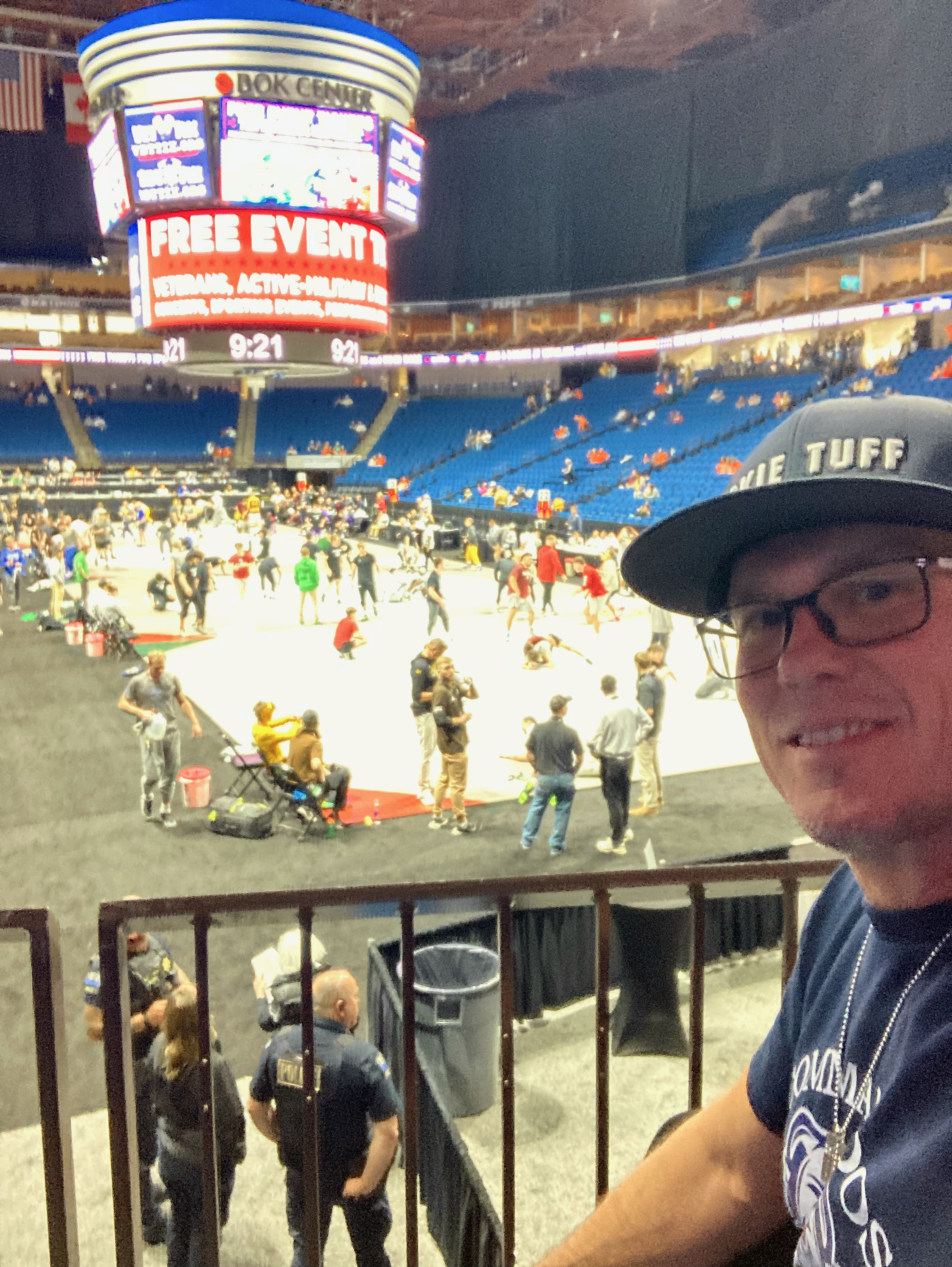 A person wearing a baseball cap and glasses, smiling, is in the foreground. In the background, there is an arena with many people walking around and sitting. The arena has a large, brightly lit digital sign and screens, and a high ceiling with murals or decorations. The seat area has blue chairs and some people are wearing orange vests.
