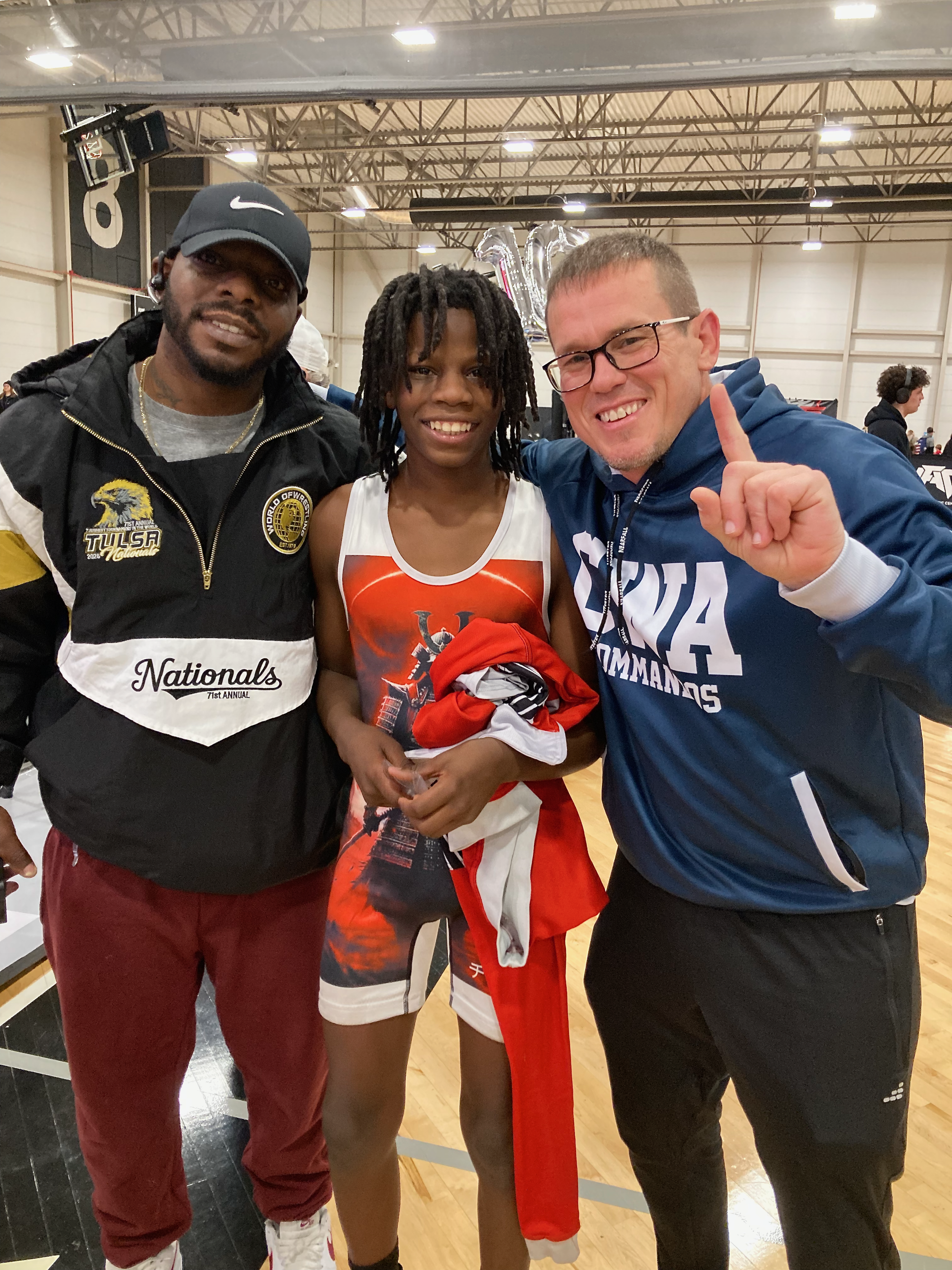 Three people standing inside a gymnasium, celebrating after a sports event. The young woman at the center has dreadlocks and is wearing a sports jersey and shorts. The man on the right wearing glasses and a navy jacket is holding up his finger, indicating number one. The man on the left wearing a Tulsas jacket, red pants, and a black Nike cap. There are large balloons in the background, spelling out the number 1, and gym equipment and people can be seen in the background.