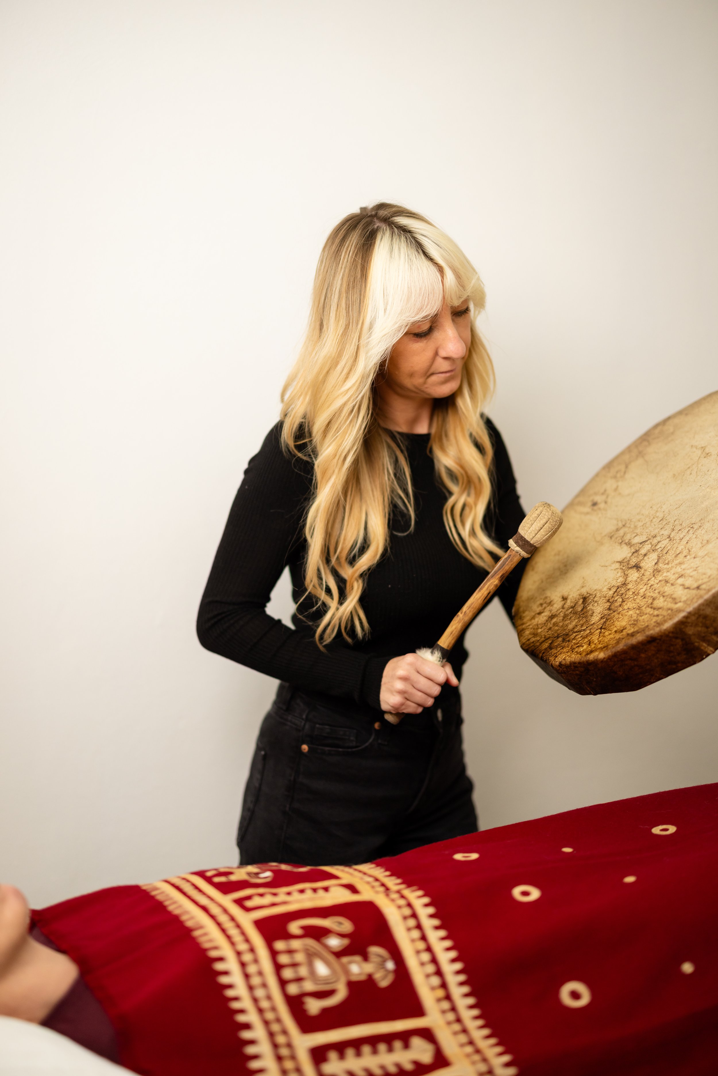 A woman with blonde hair playing a large drum, holding a drumstick in her right hand, standing in front of a white wall, with a person lying on a table with a red cloth in front of them.