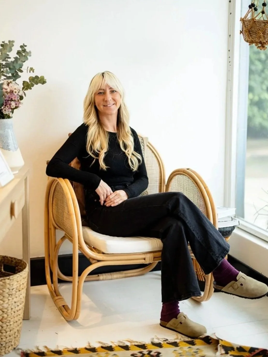 A woman with long blonde hair sitting on a rattan chair in a bright room near a window, smiling at the camera.