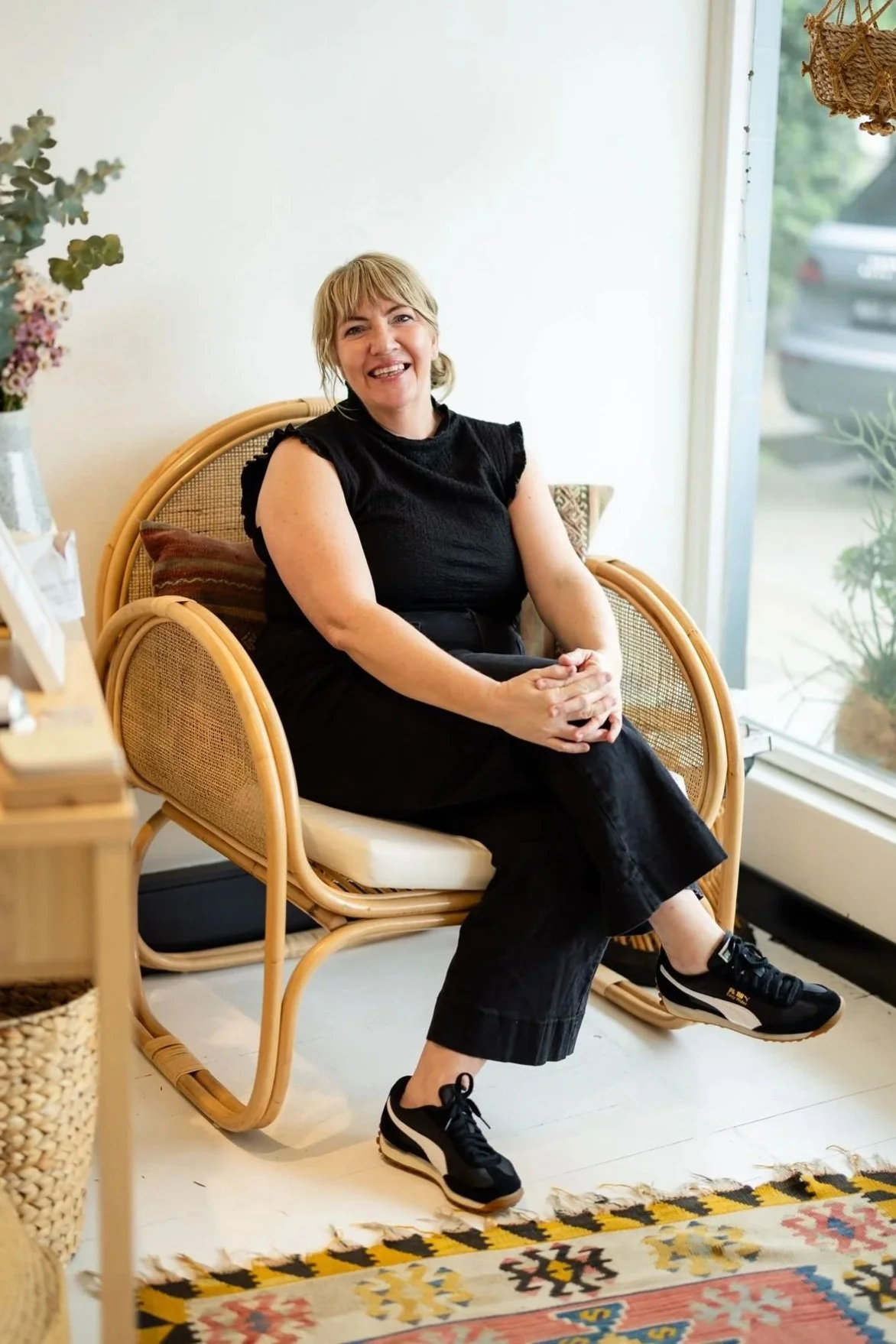 A woman with blonde hair, wearing a black sleeveless top, black pants, and black sneakers, sitting on a rattan chair and smiling in a well-lit room with a white wall, a window, and a colourful patterned rug.
