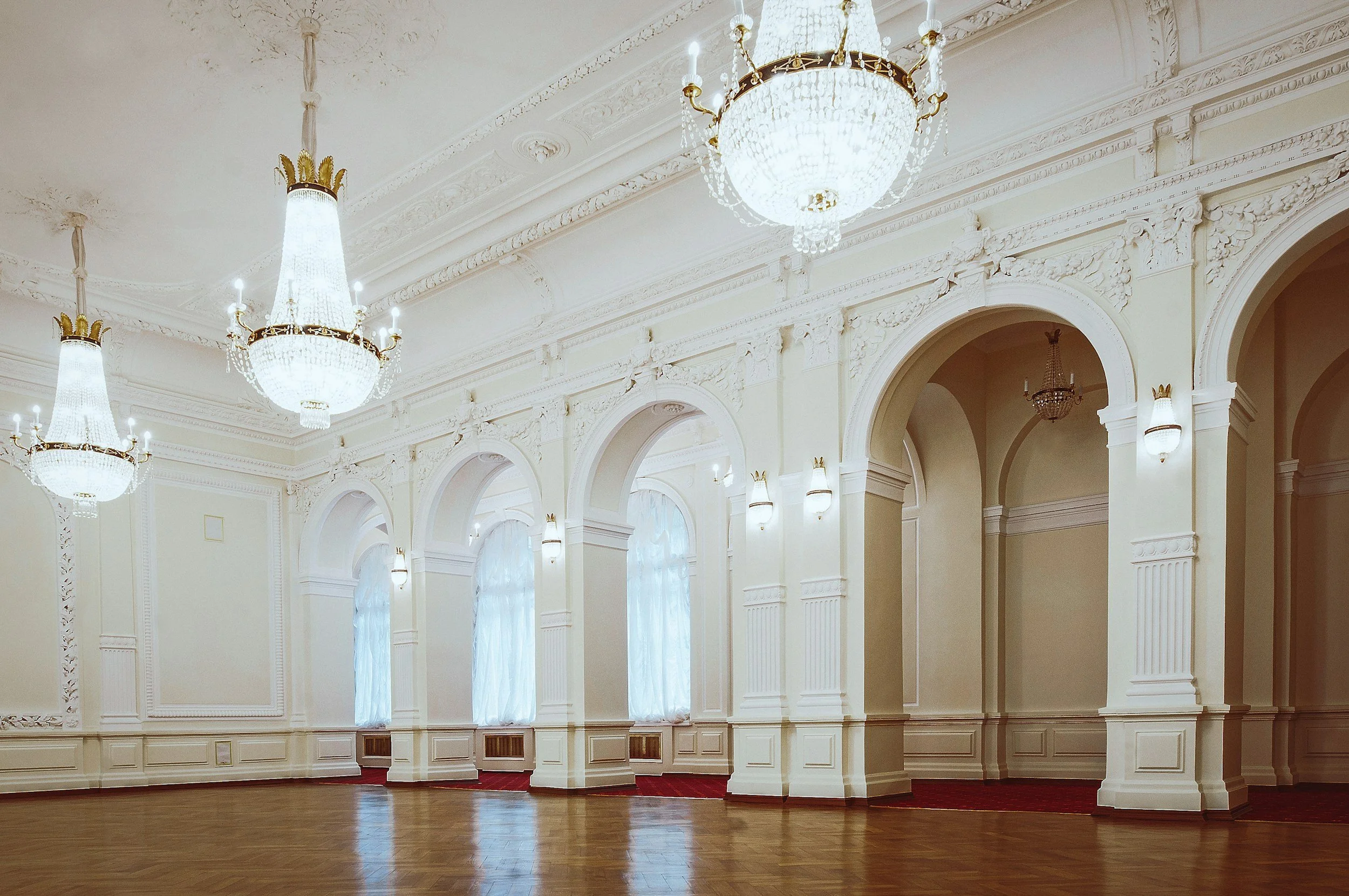 Elegant ballroom with ornate white walls, large arched windows with sheer curtains, and several grand chandeliers hanging from a decorated ceiling.