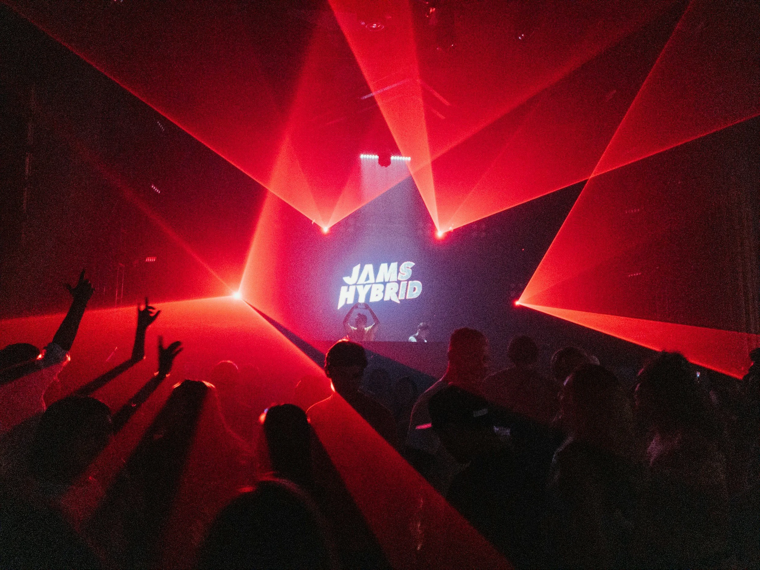 A concert scene with red laser lights and a crowd dancing, with a screen displaying the text 'JAMS HYBRID' in the background.