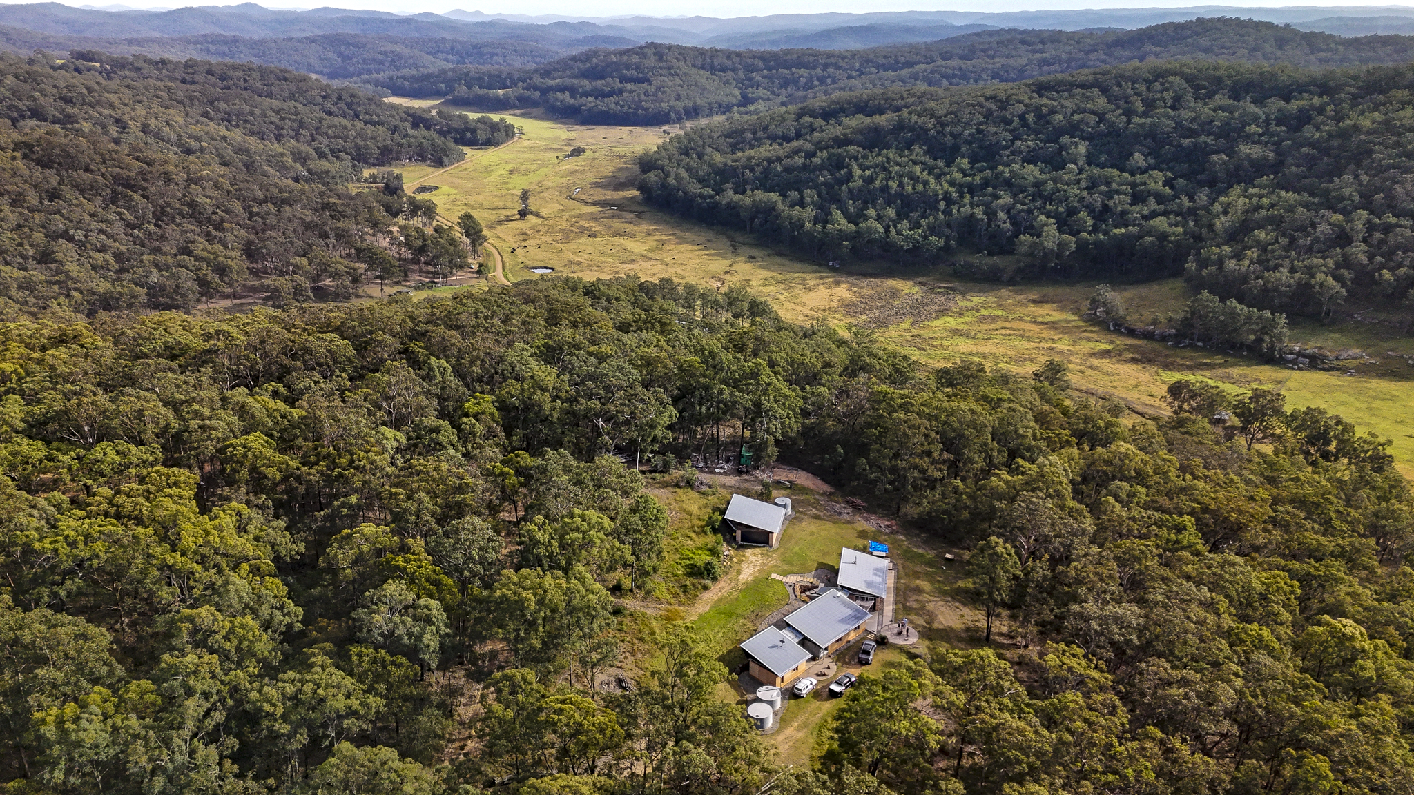 An aerial view of a forested valley with a small clearing containing a few structures, surrounded by dense trees and rolling hills in the distance.