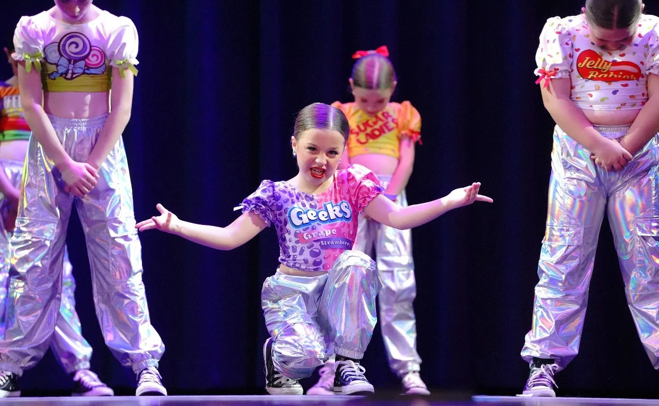 Young girls performing a dance on stage with holographic pants and bright, colorful shirts.