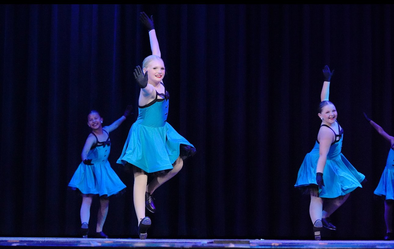 Four young girls in blue dance costumes performing on stage with black curtains, mid-dance with raised arms and smiling.