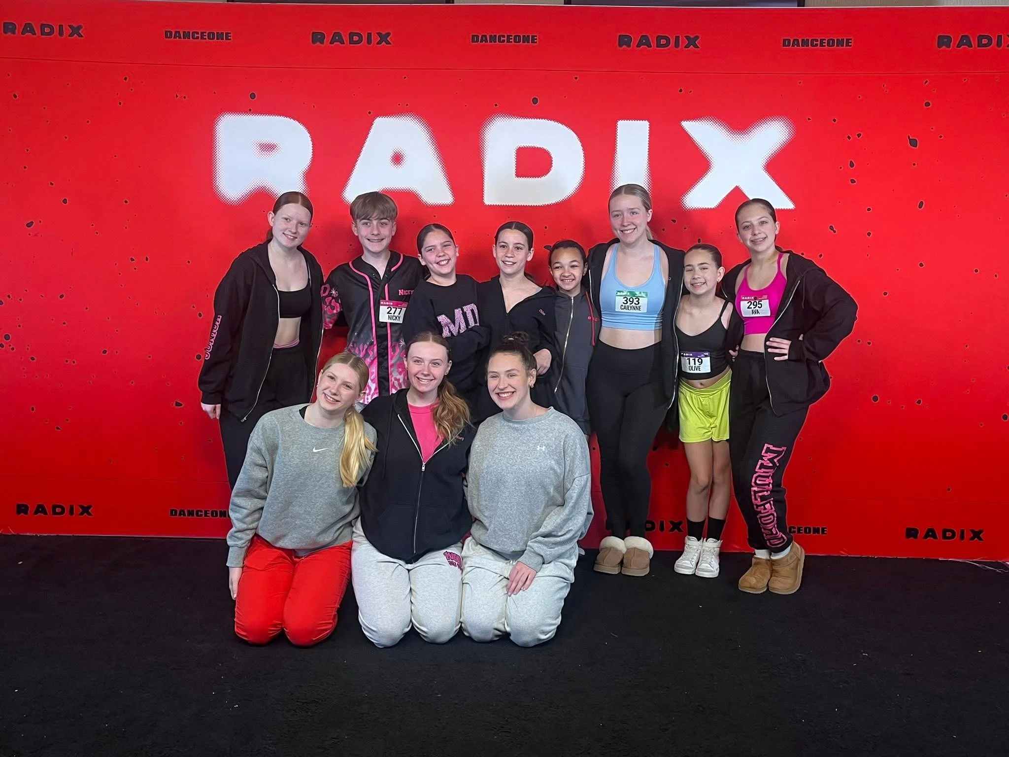A group of ten young female athletes in sportswear posing together in front of a red backdrop with the words "RADIX" and "DANCEONE" printed on it.