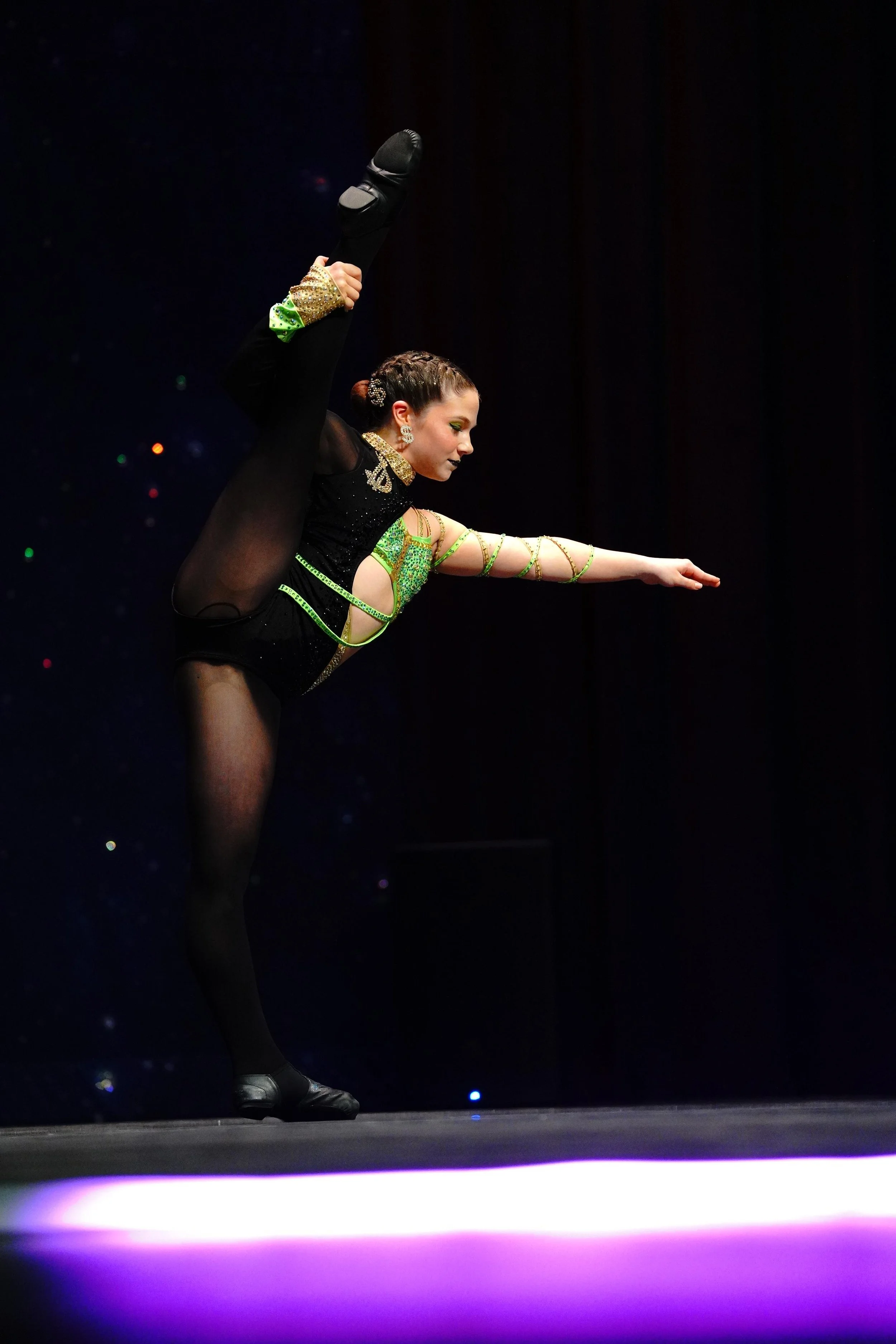 A female gymnastPerforming a leg lift on stage with a dark background.