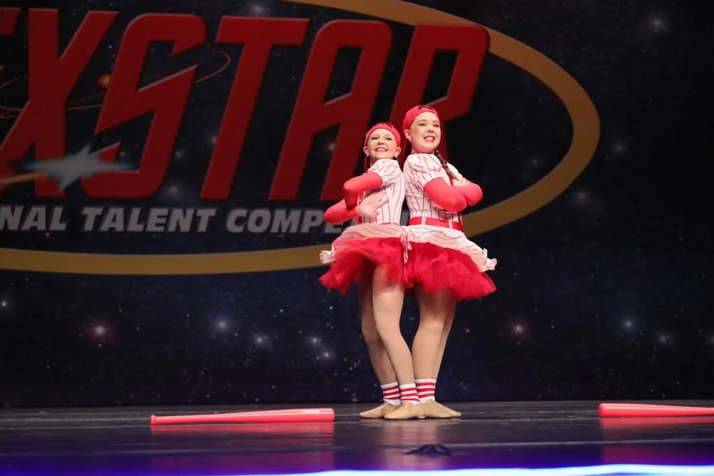 Two young girls dressed in coordinated red and white costumes, standing back-to-back with arms crossed, smiling on stage at a talent competition with a large backdrop displaying the word 'XSTAR' and a space-themed background.