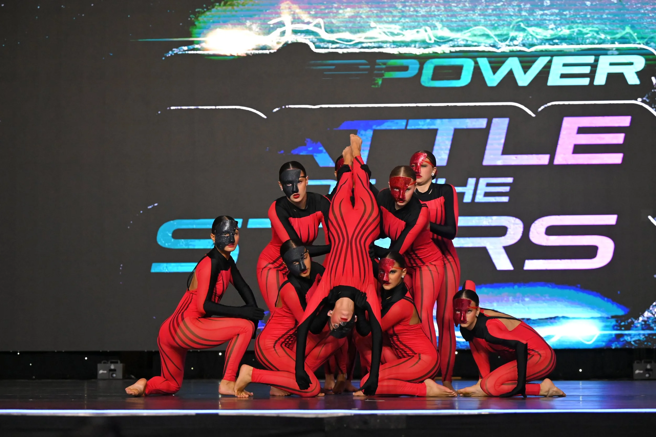 A group of nine dancers in coordinated red and black costumes poses on stage during a dance performance with a digital backdrop displaying the words 'Power', 'Battle', and 'Stars'.