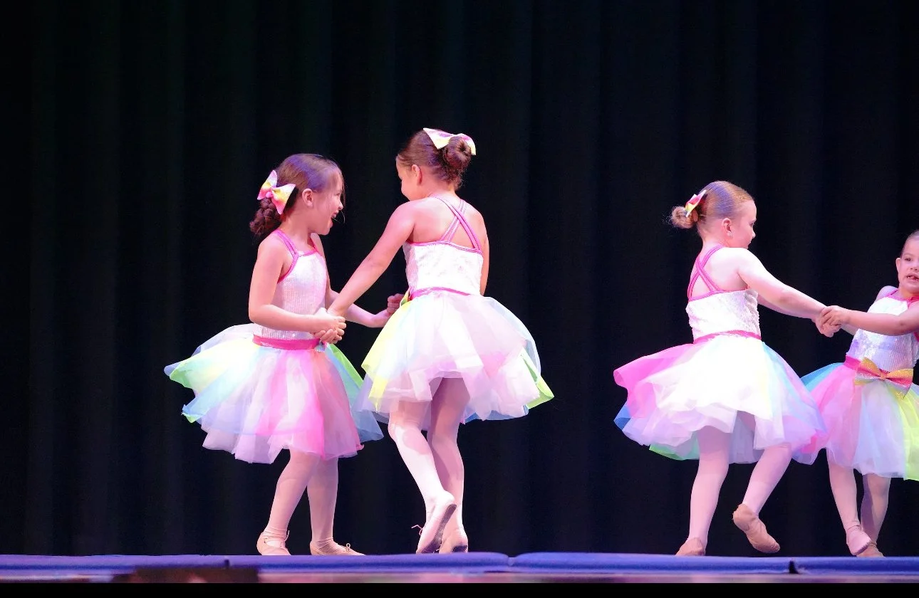 Four young girls in colorful tutu dresses and ballet shoes holding hands and dancing on stage with a dark curtain backdrop.