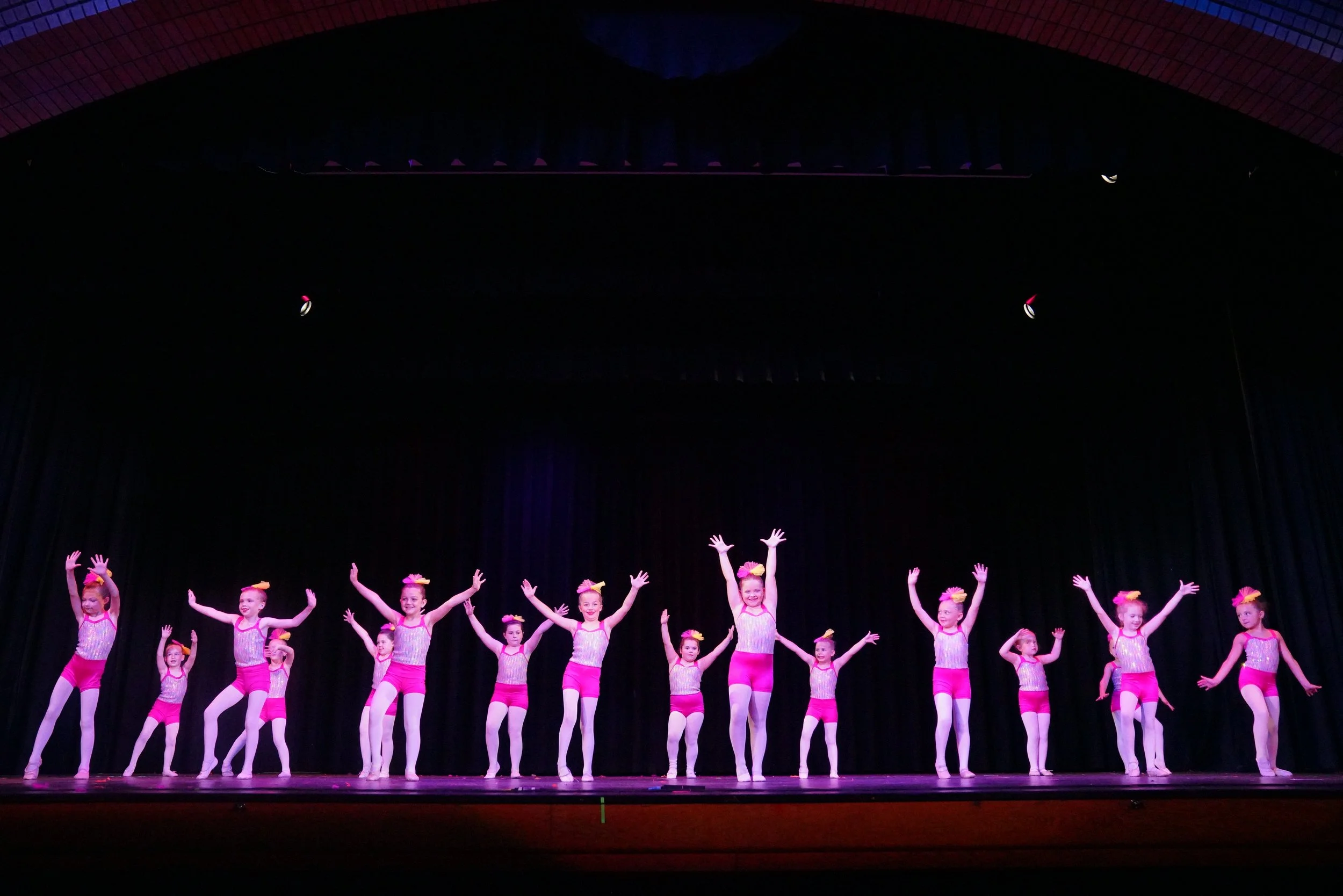Young girls performing a dance on stage, wearing pink costumes with yellow bows and white tights.