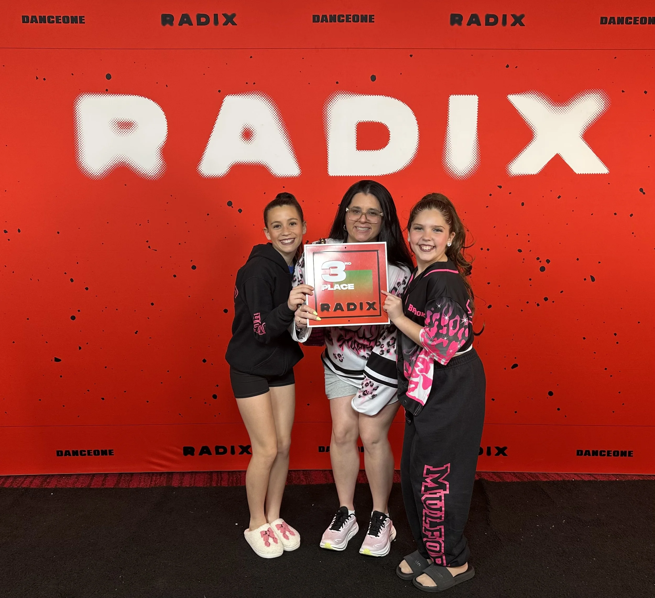 Three smiling girls standing in front of a red background with the word 'RADIX' in bold white letters. They are holding a red and black certificate that says '3rd Place RADIX' and 'DANCEONE'. The girls are wearing casual and dance-themed clothing, including shorts, leggings, and a hoodie.