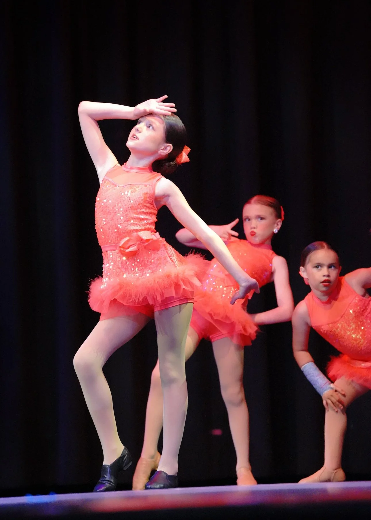 Three young girls in orange costumes performing a dance on stage with black curtains behind them.