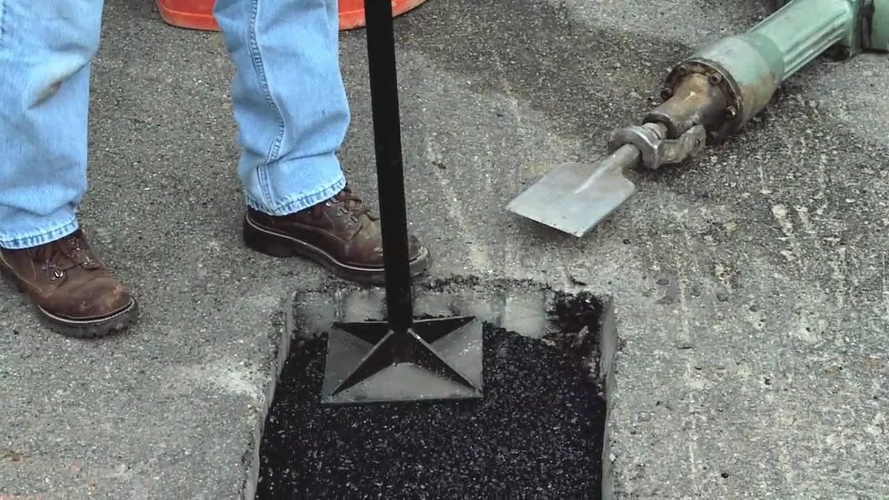 Worker pouring asphalt into a hole in the pavement using a large machine, wearing jeans and brown boots.