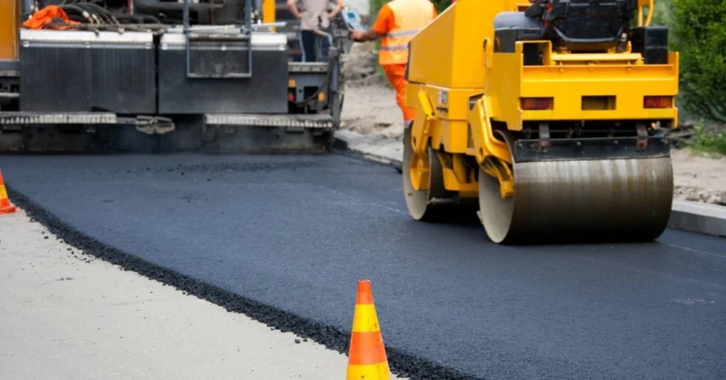 Construction workers paving a road with asphalt, using heavy machinery and orange traffic cones.