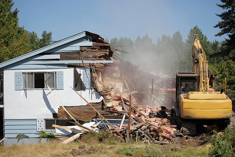 A yellow excavator demolishing a house with part of the structure falling apart and dust in the air.