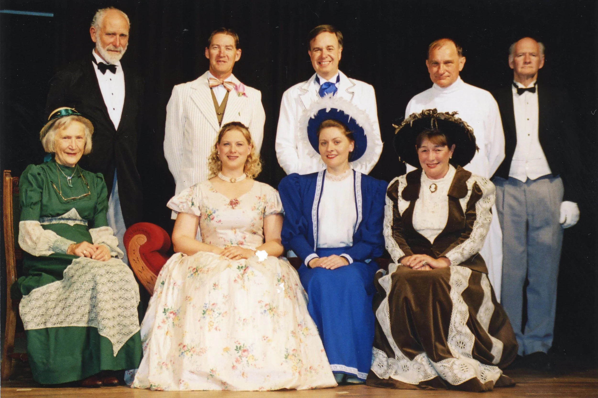 Group of nine people dressed in vintage, historical costumes, posing for a photo on stage.