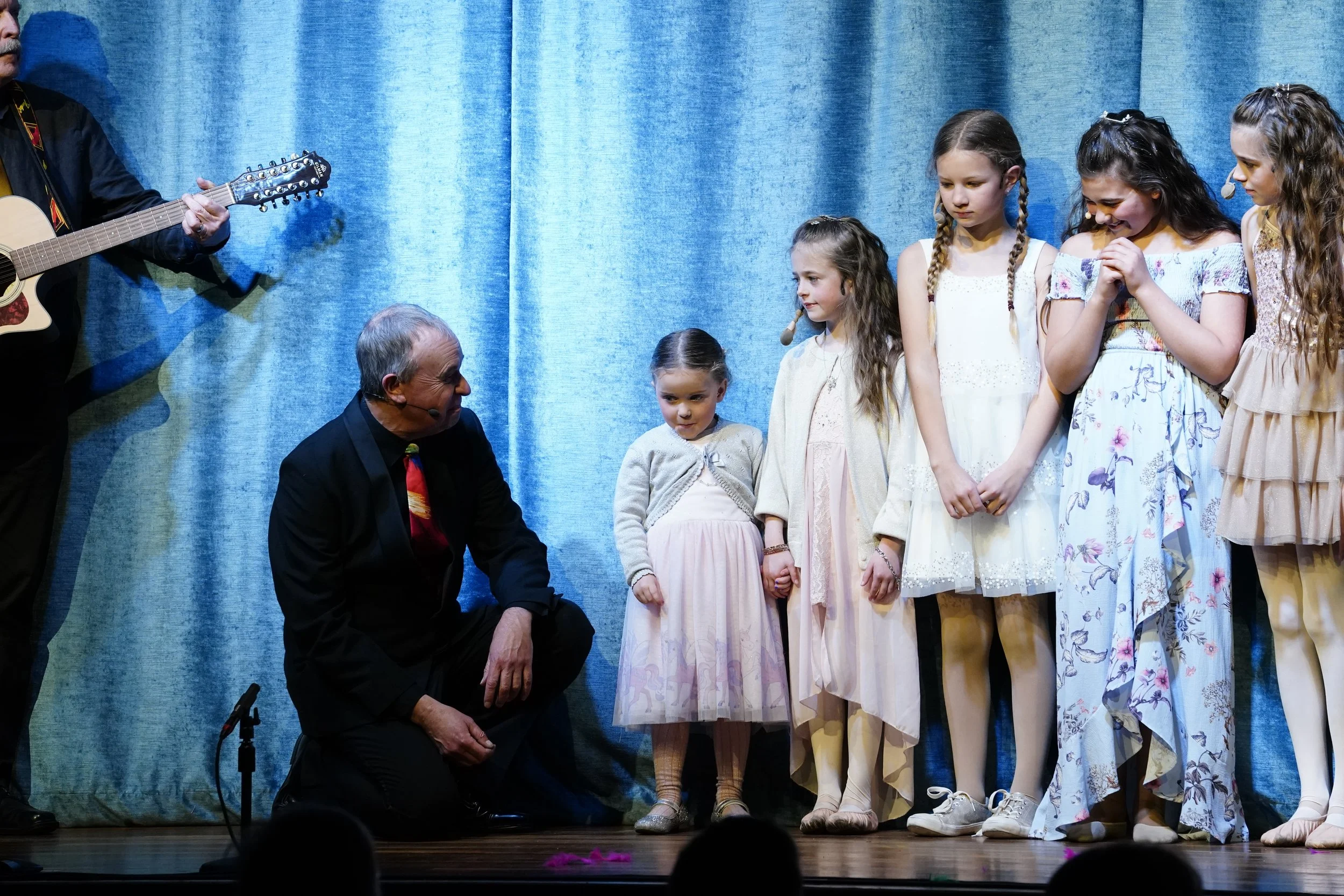 Children and adults on stage, some holding hands, with a man playing guitar on the left, in front of a blue curtain.