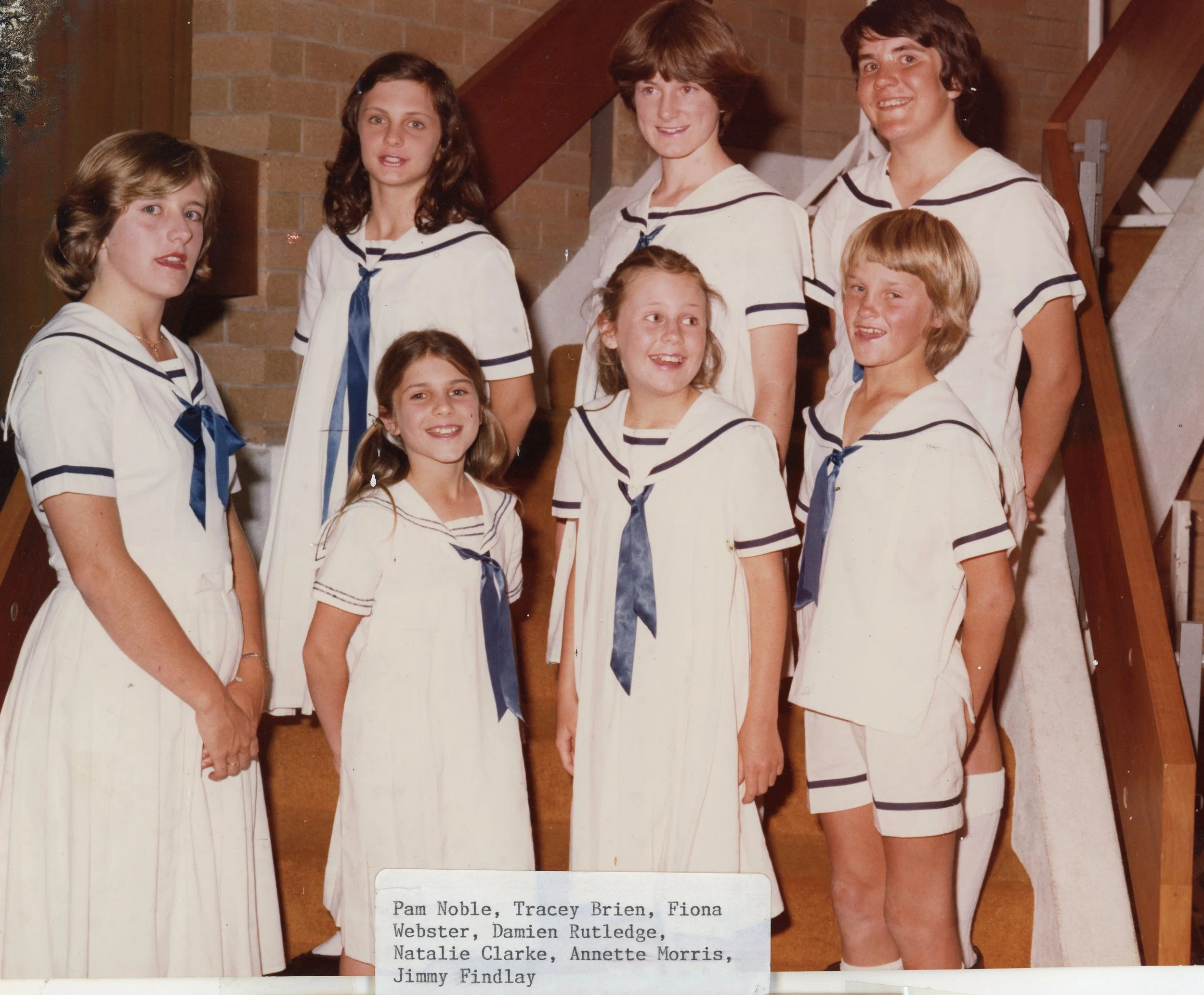 A group of nine young girls dressed in vintage white sailor dresses with navy blue accents, standing on a staircase indoors. They are smiling and posing for a photo.