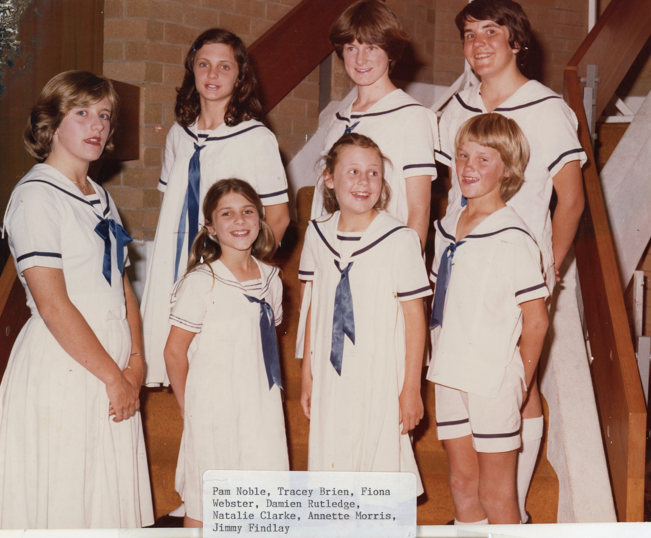 Photograph of eight young girls in vintage sailor-style dresses standing on a staircase indoors, smiling at the camera.