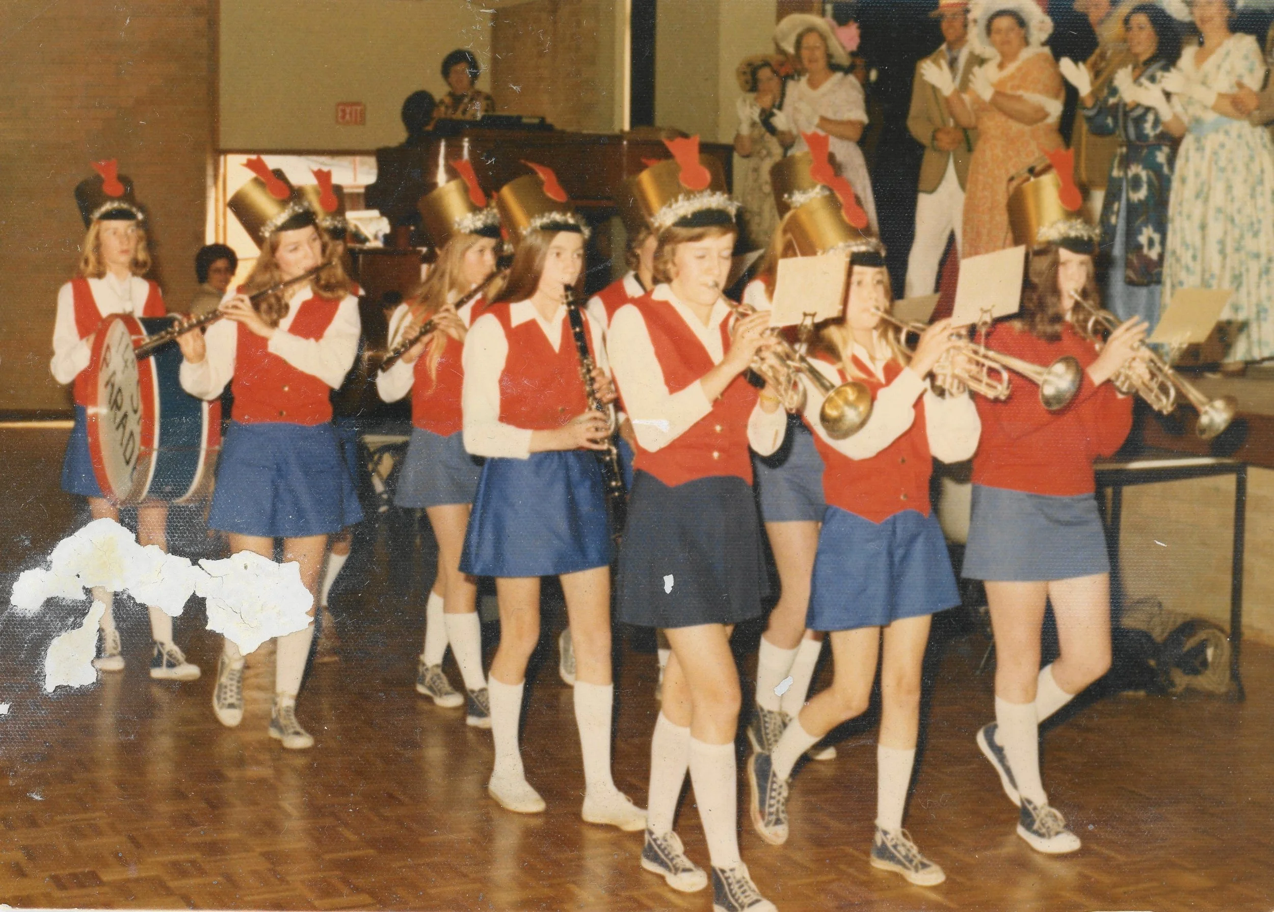 A group of young girls dressed in matching red, white, and blue outfits performing with musical instruments, including trumpets, xylophones, and clarinets, during a holiday or Christmas-themed event. They are walking in a line on a wooden floor in fr