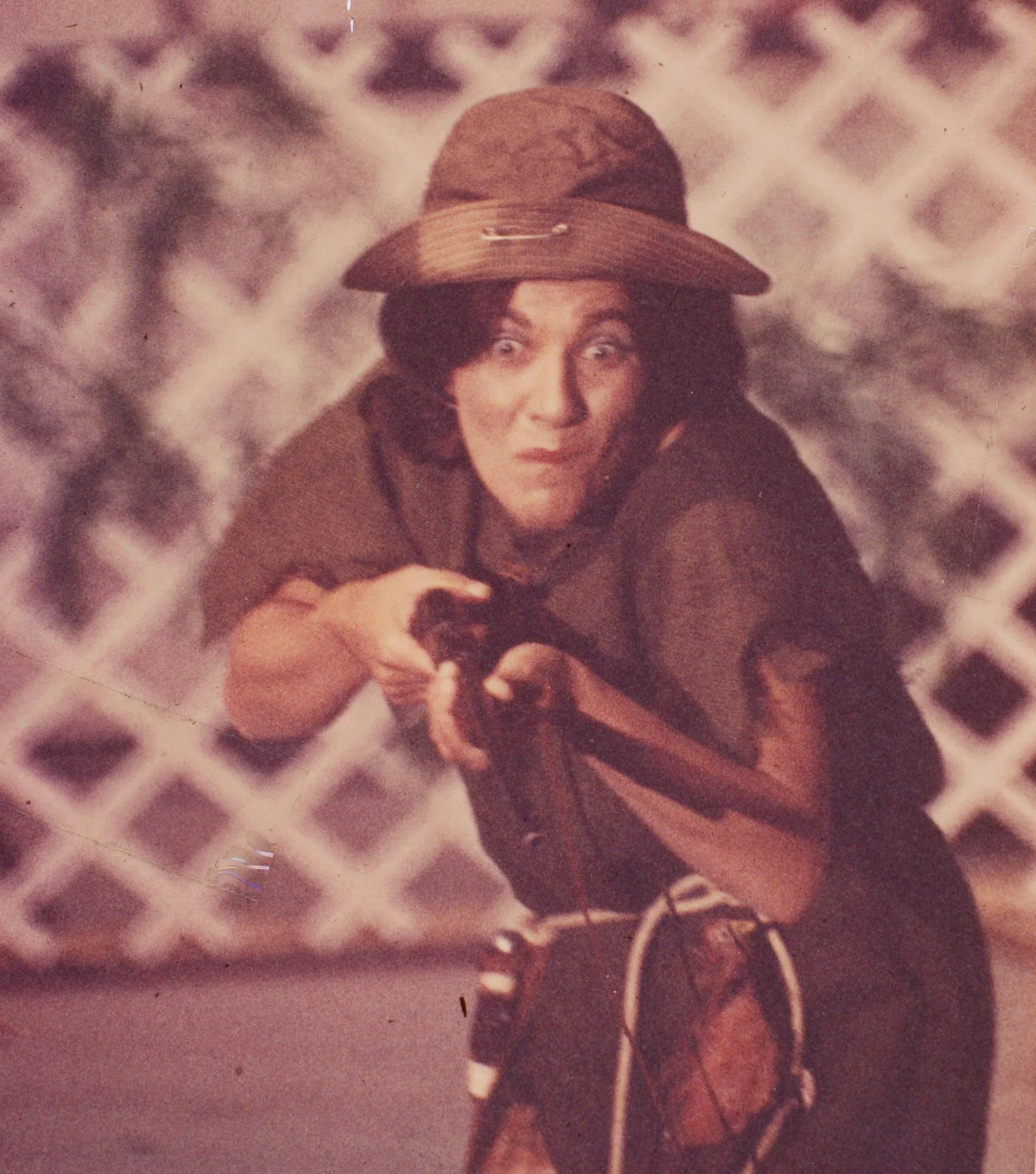 A woman wearing a wide-brimmed hat and brown clothing, holding a black and white puppy, with a white lattice fence background.