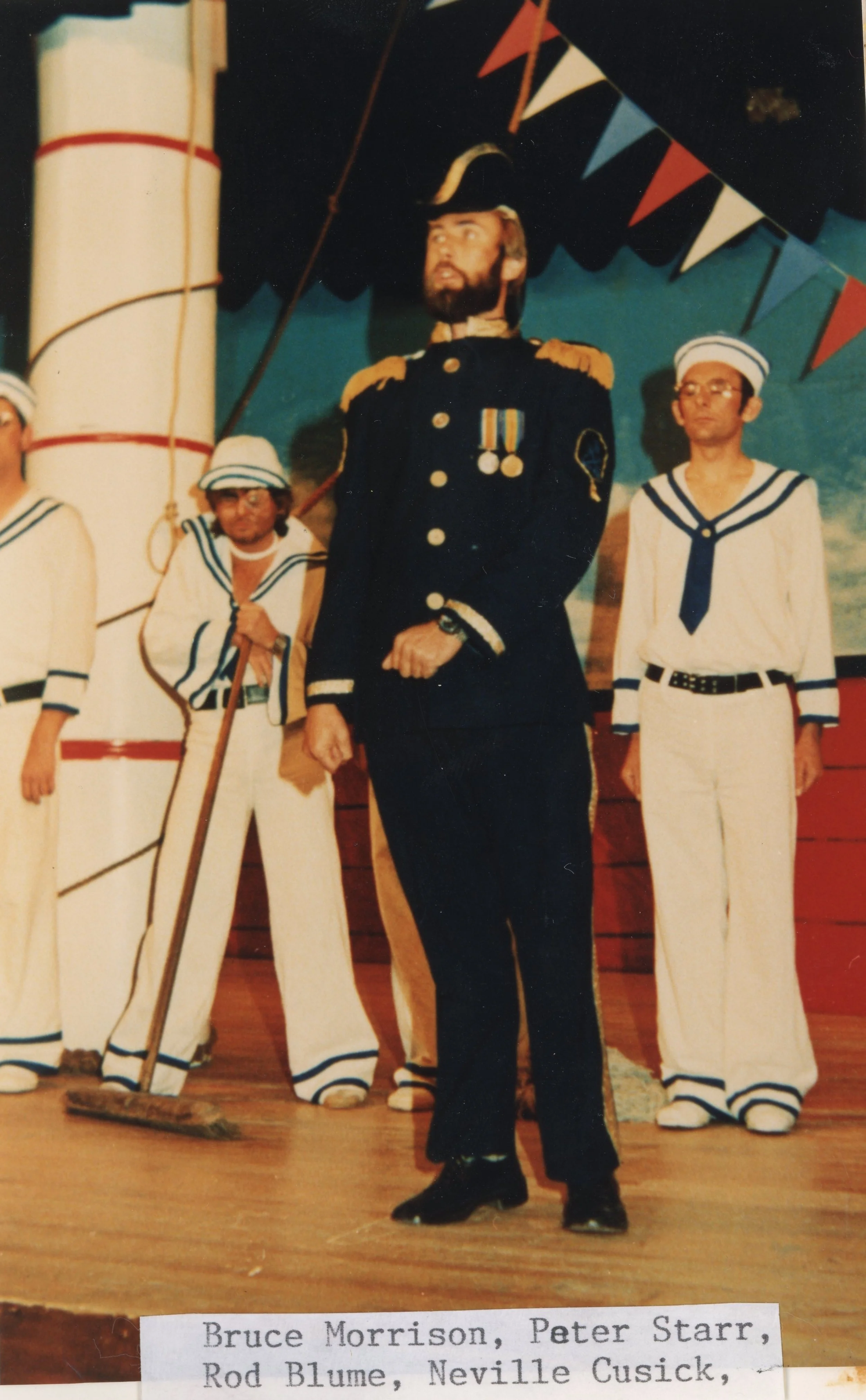 Stage performance featuring individuals dressed in nautical and military costumes, including a man in a military uniform with medals, surrounded by others in sailor outfits, on a decorated stage.