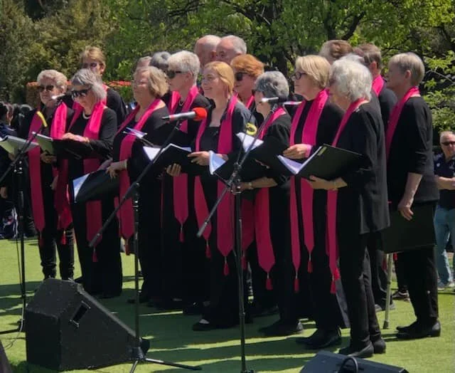 Cowra Theatre Company A choir of elderly women wearing black outfits with pink scarves, standing outdoors on a grassy area, singing into microphones during a performance.