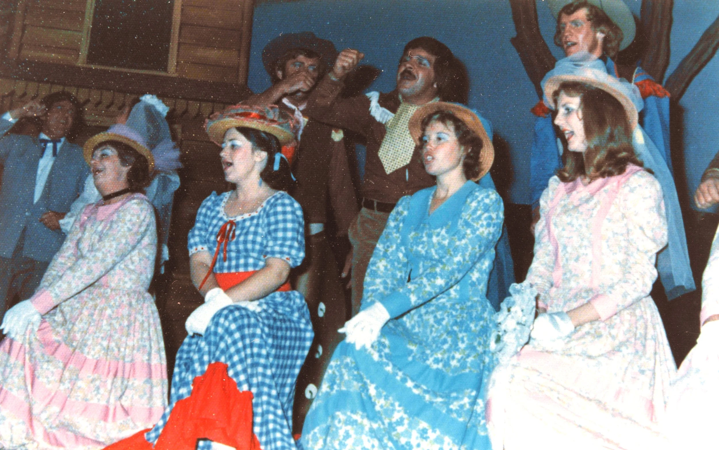 Group of actors on stage, dressed in old-fashioned Western dresses and suits, performing in a musical or play, with painted background and wooden set pieces.