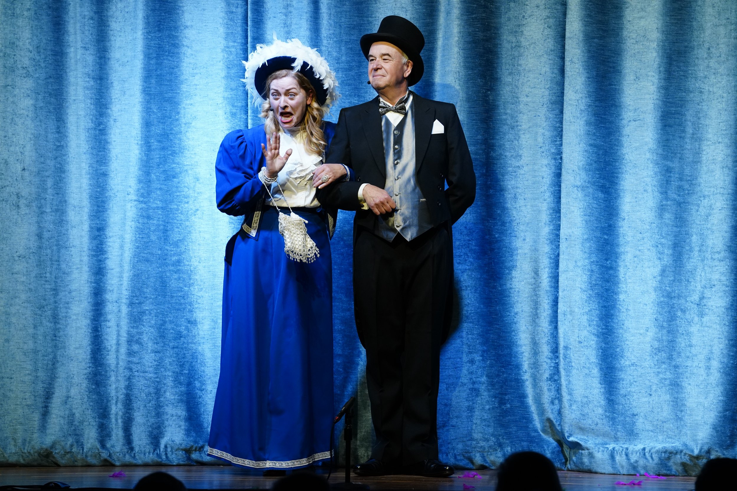 Actors performing on stage with blue curtain backdrop, dressed in period costumes; woman in blue dress and feathered hat, man in black tuxedo with top hat.