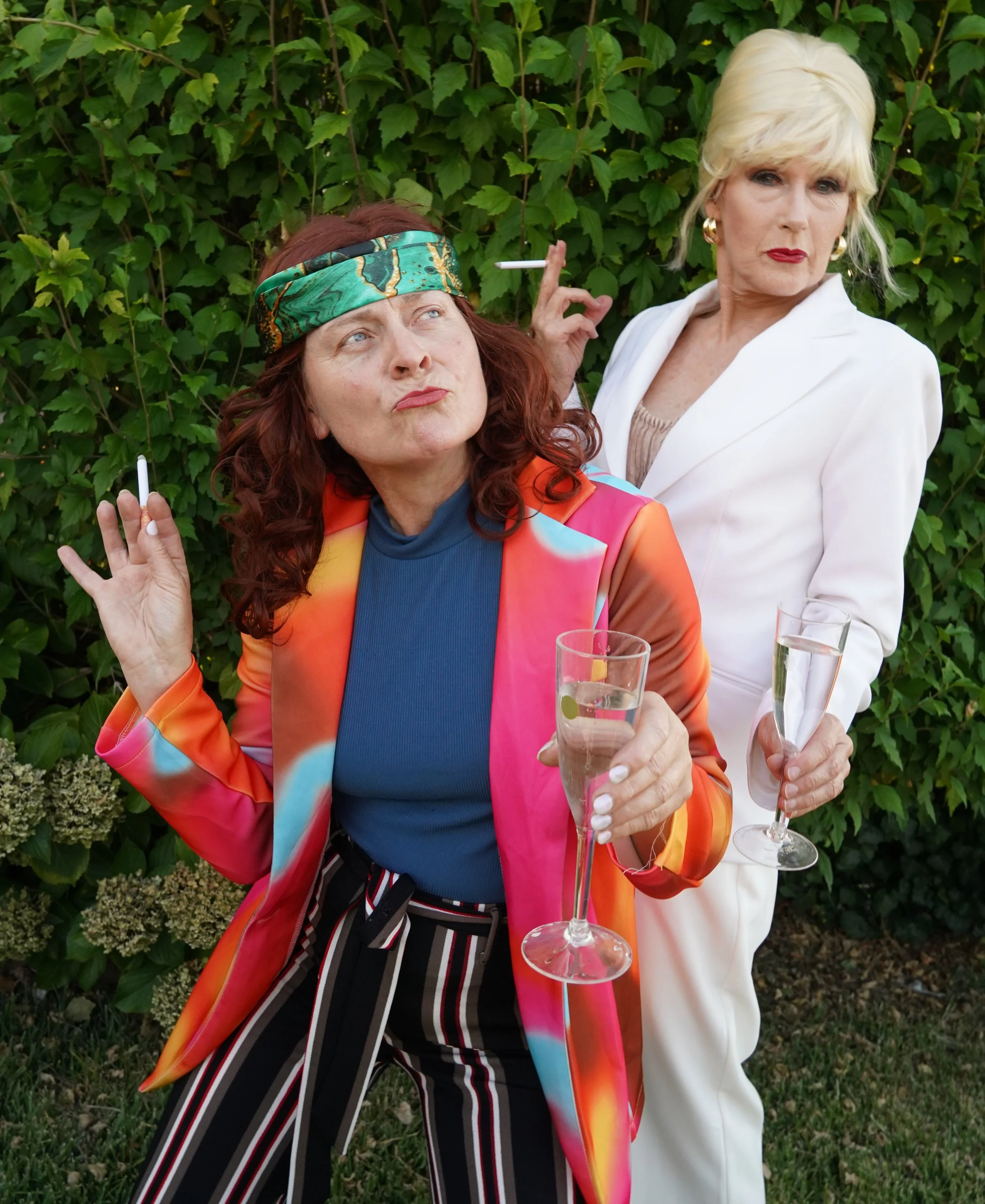 Cowra Theatre Company Two women outdoors holding glasses of champagne and cigarettes, one wearing a colorful blazer and striped pants, the other dressed in a white suit, with a green leafy background.