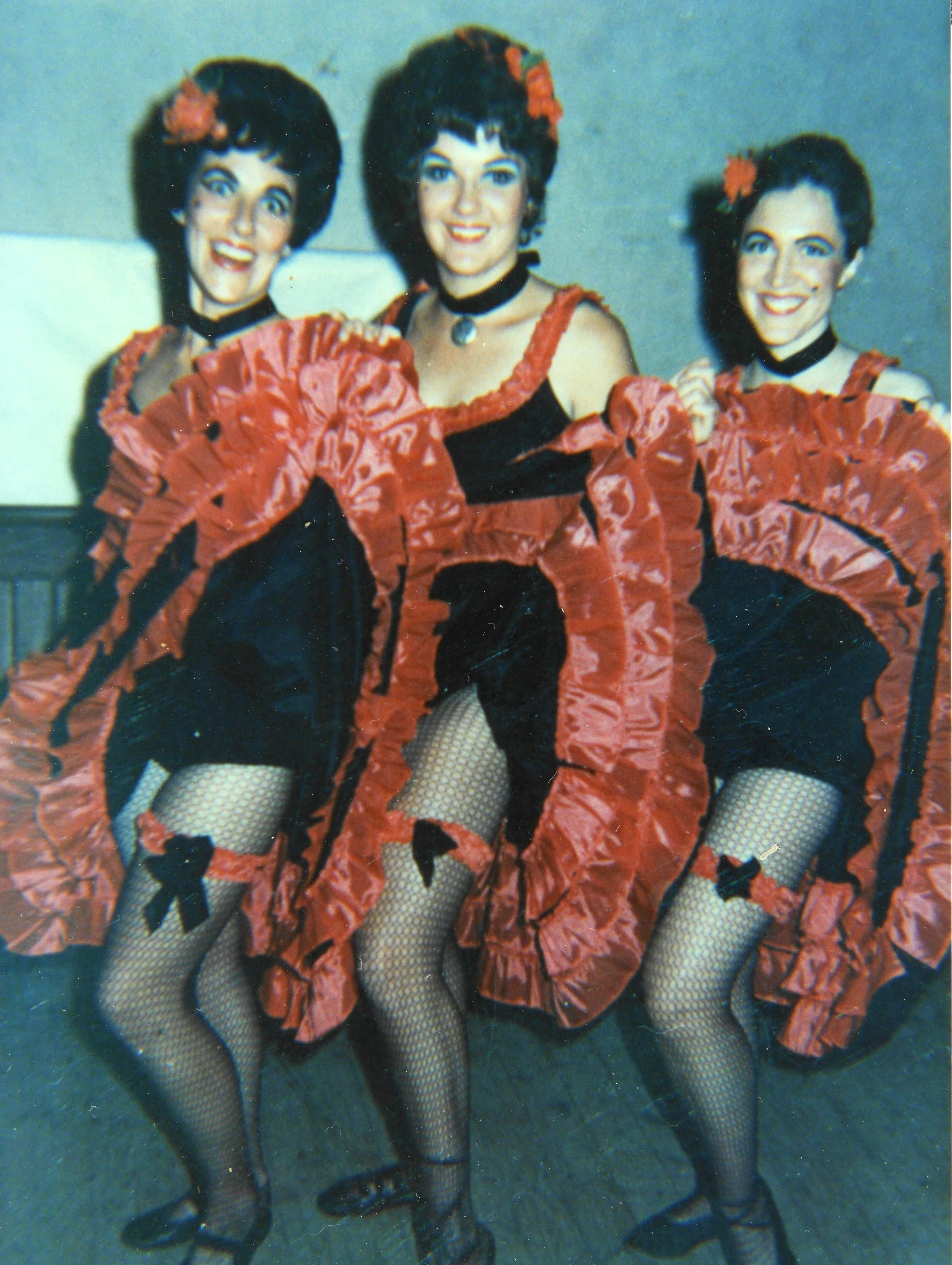 Three women dressed in black and red flamenco-style costumes with ruffled skirts, fishnet stockings, and flower accessories in their hair, performing a dance.