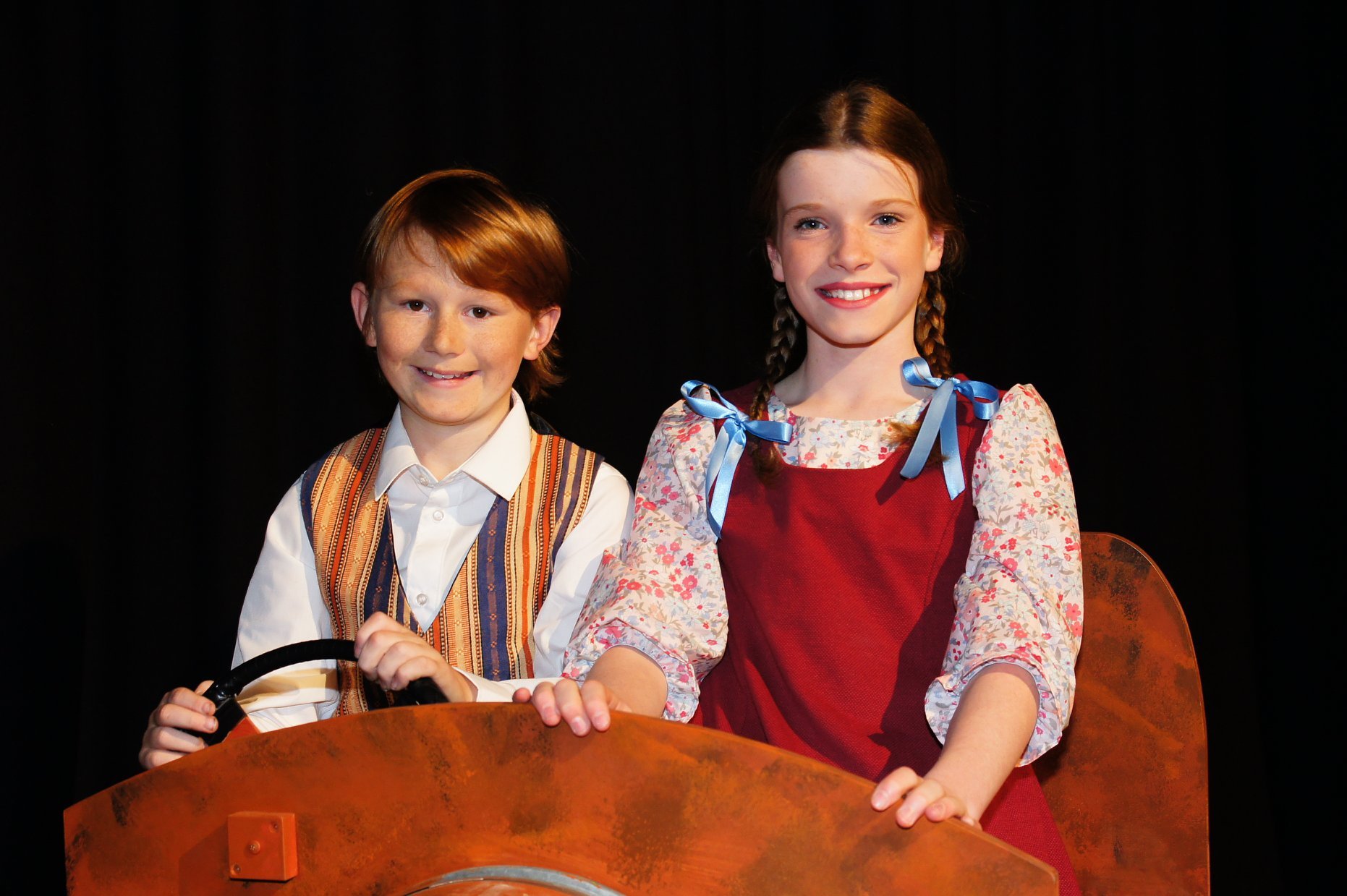 Cowra Theatre Company Cowra Theatre Company Two children on a stage, a boy and a girl, smiling, holding a steering wheel, dressed in costumes, with a black background.