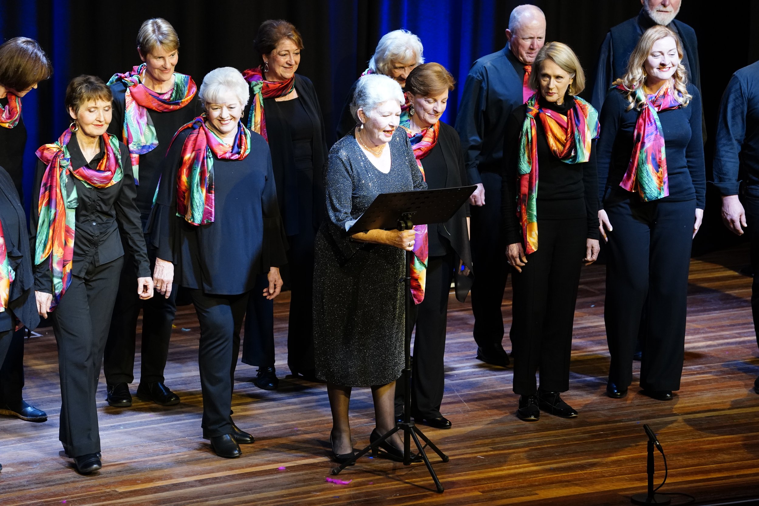 A group of older adults standing on a stage, dressed in black with colorful scarves, with one woman standing at a music stand reading or singing during a performance.
