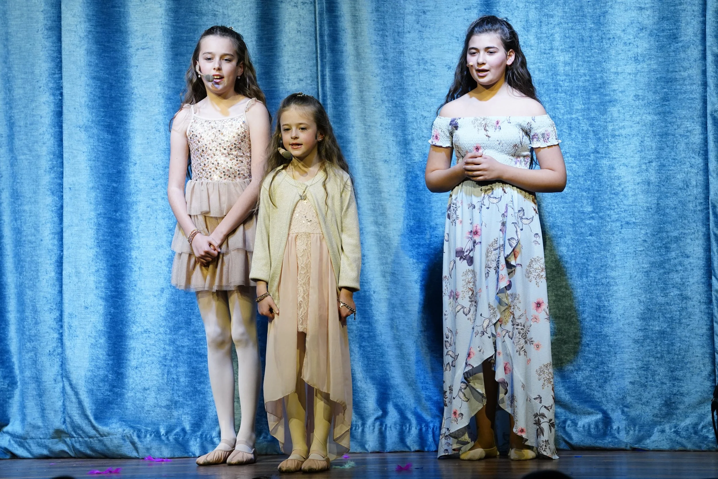 Four young girls in dresses standing on a stage with a blue curtain background, wearing microphones, likely in a performance or recital.