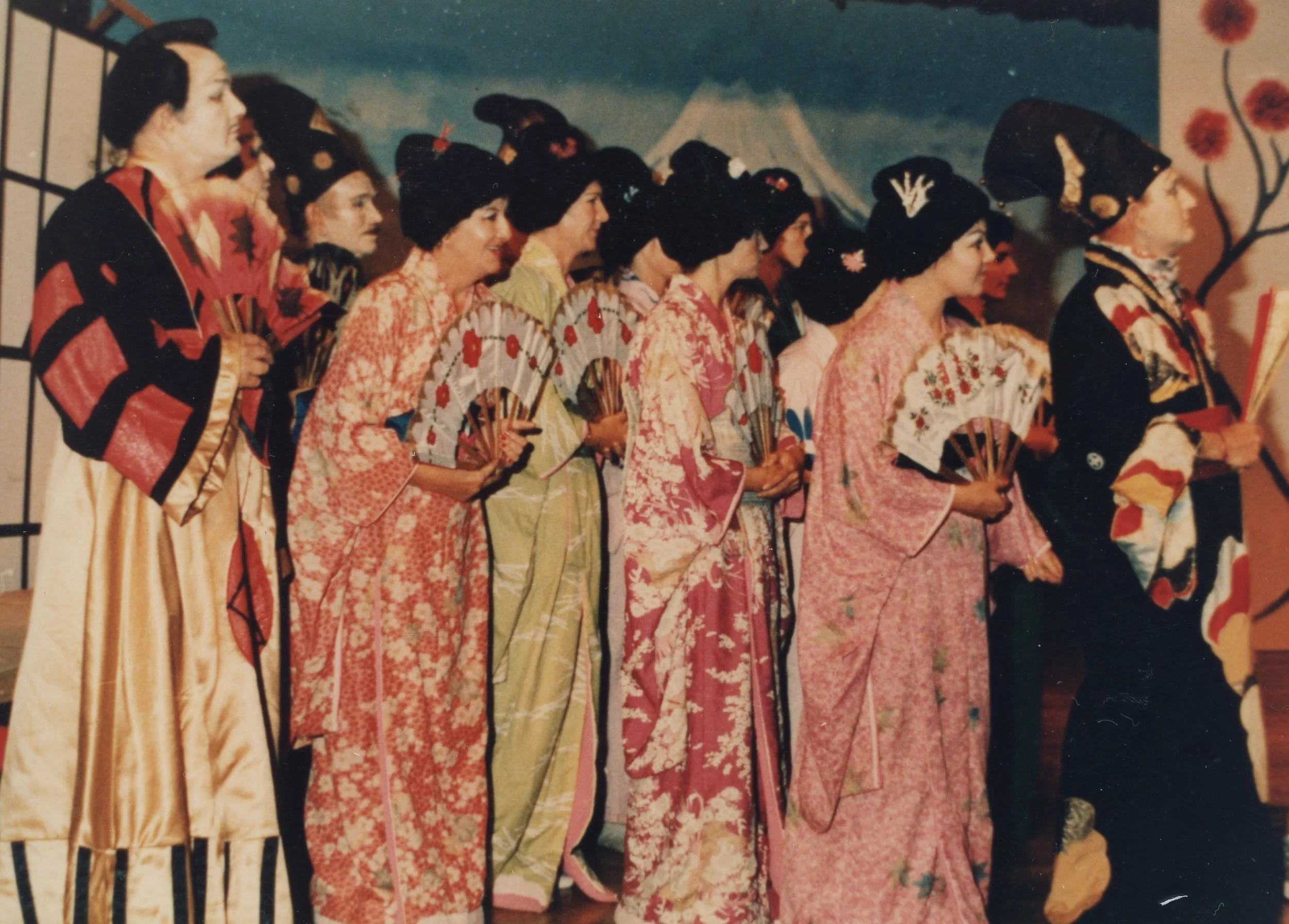 Women and a man dressed in traditional Japanese kimonos with fans, standing in a line, in front of a painted backdrop with Mount Fuji and a cherry blossom tree.