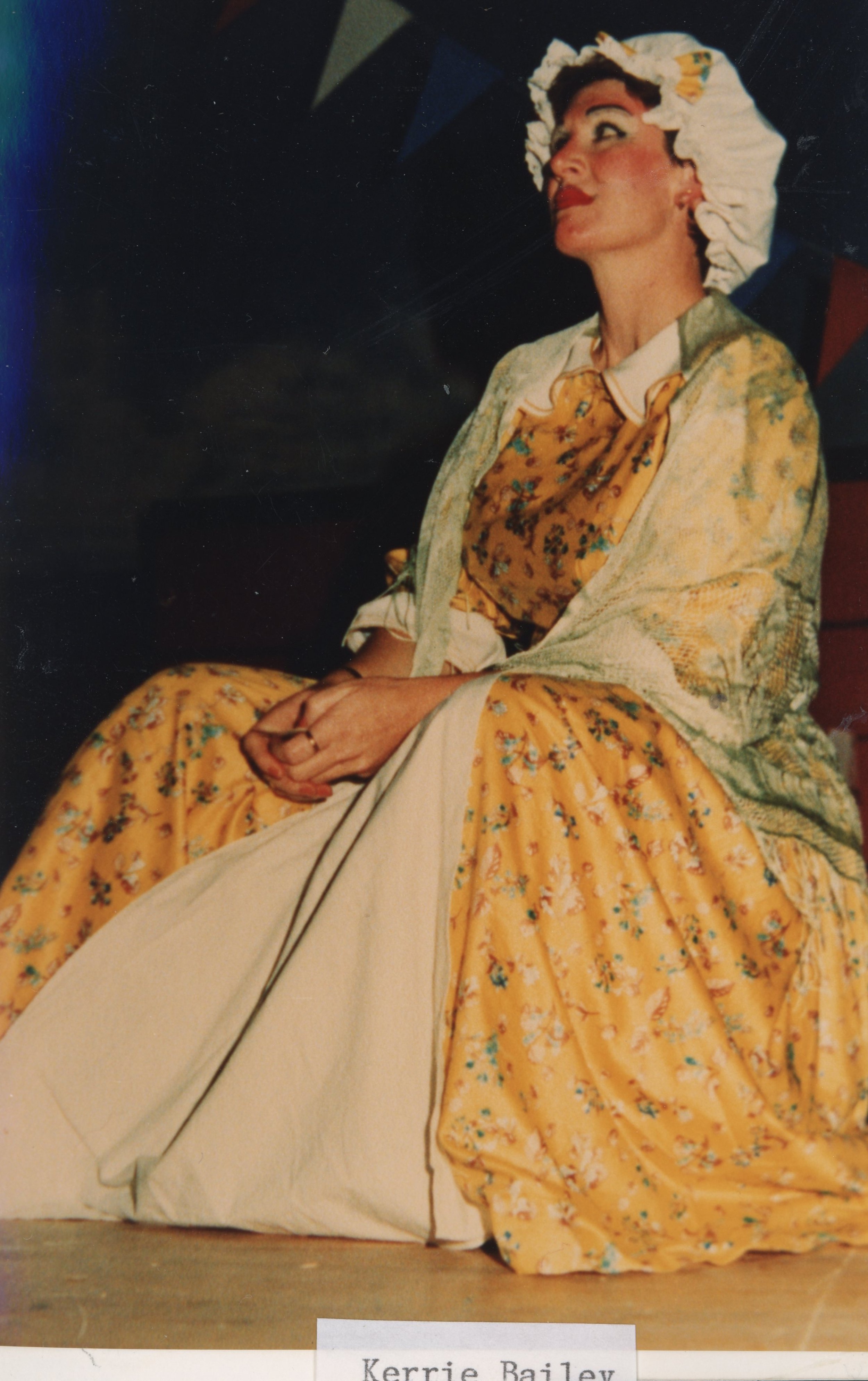 A woman dressed in a vintage costume, sitting on stage with her hands clasped in her lap, looking to her left. She wears a yellow floral dress, a light green cardigan, and a white bonnet with ruffles.